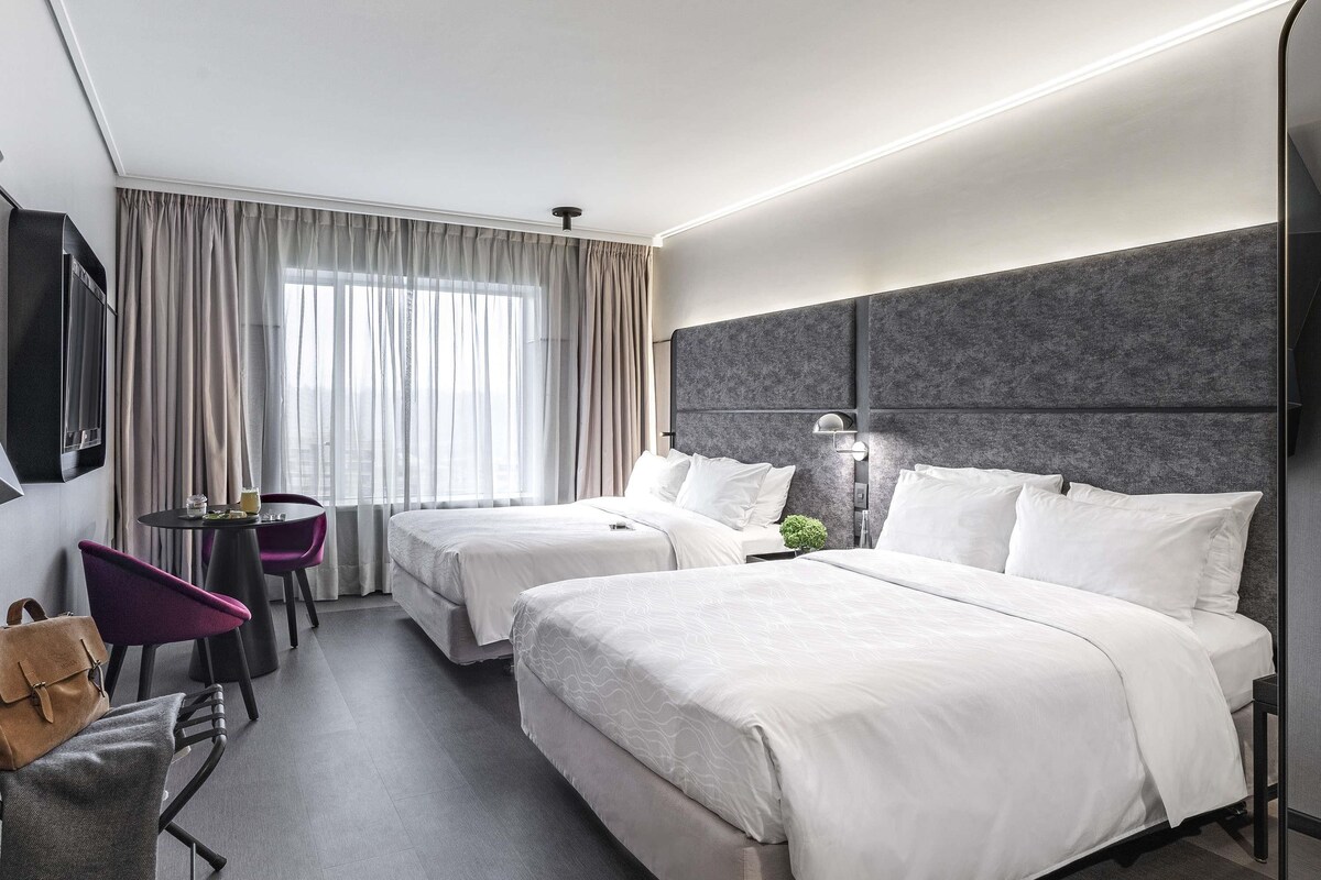 Habitación de hotel con dos camas, un televisor montado en la pared, una pequeña mesa con sillas y vistas a la ciudad a través de amplios ventanales.