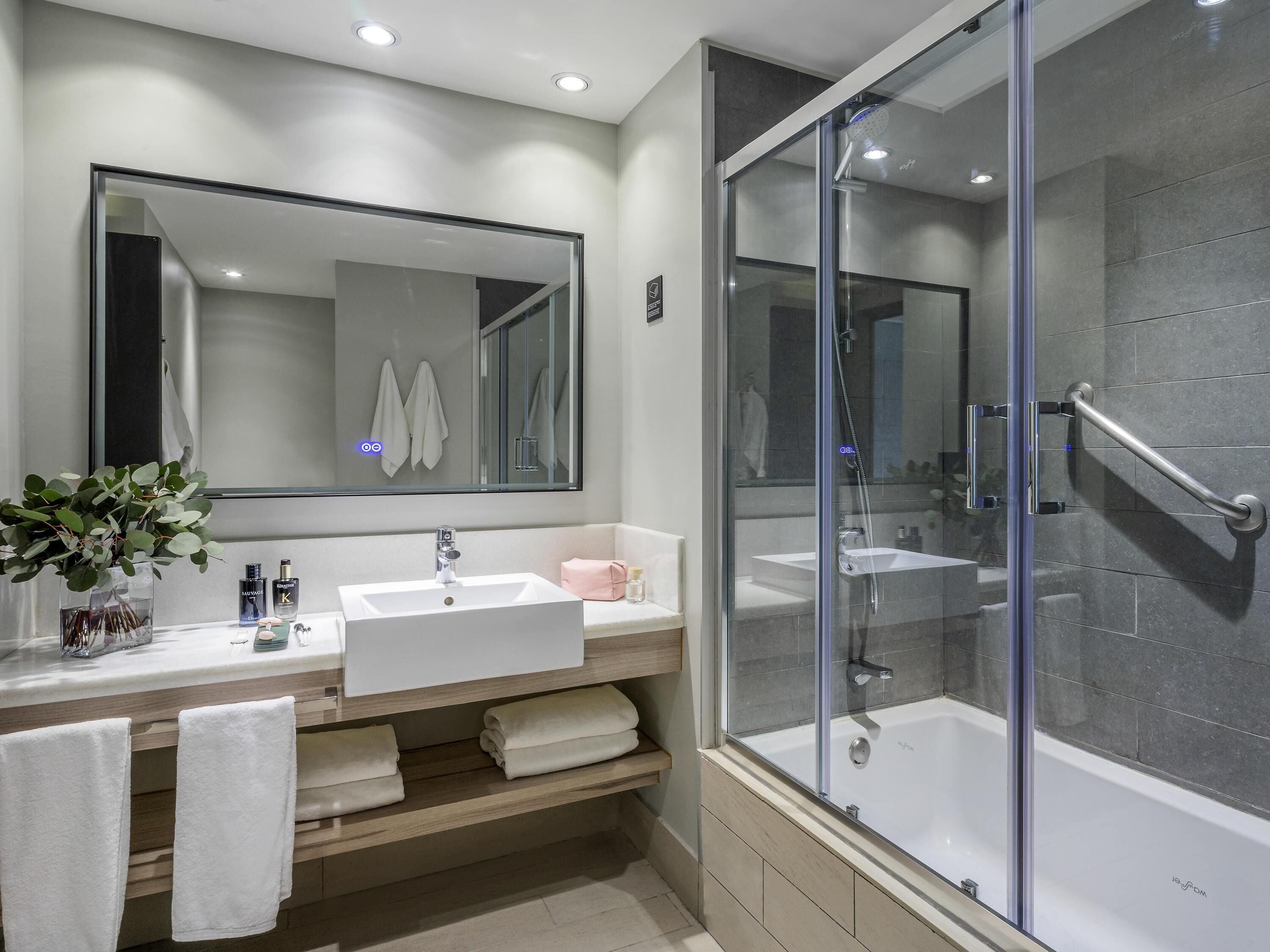 Un baño moderno con un espejo grande, un lavabo blanco, una ducha con mampara de vidrio y un tocador de madera.
