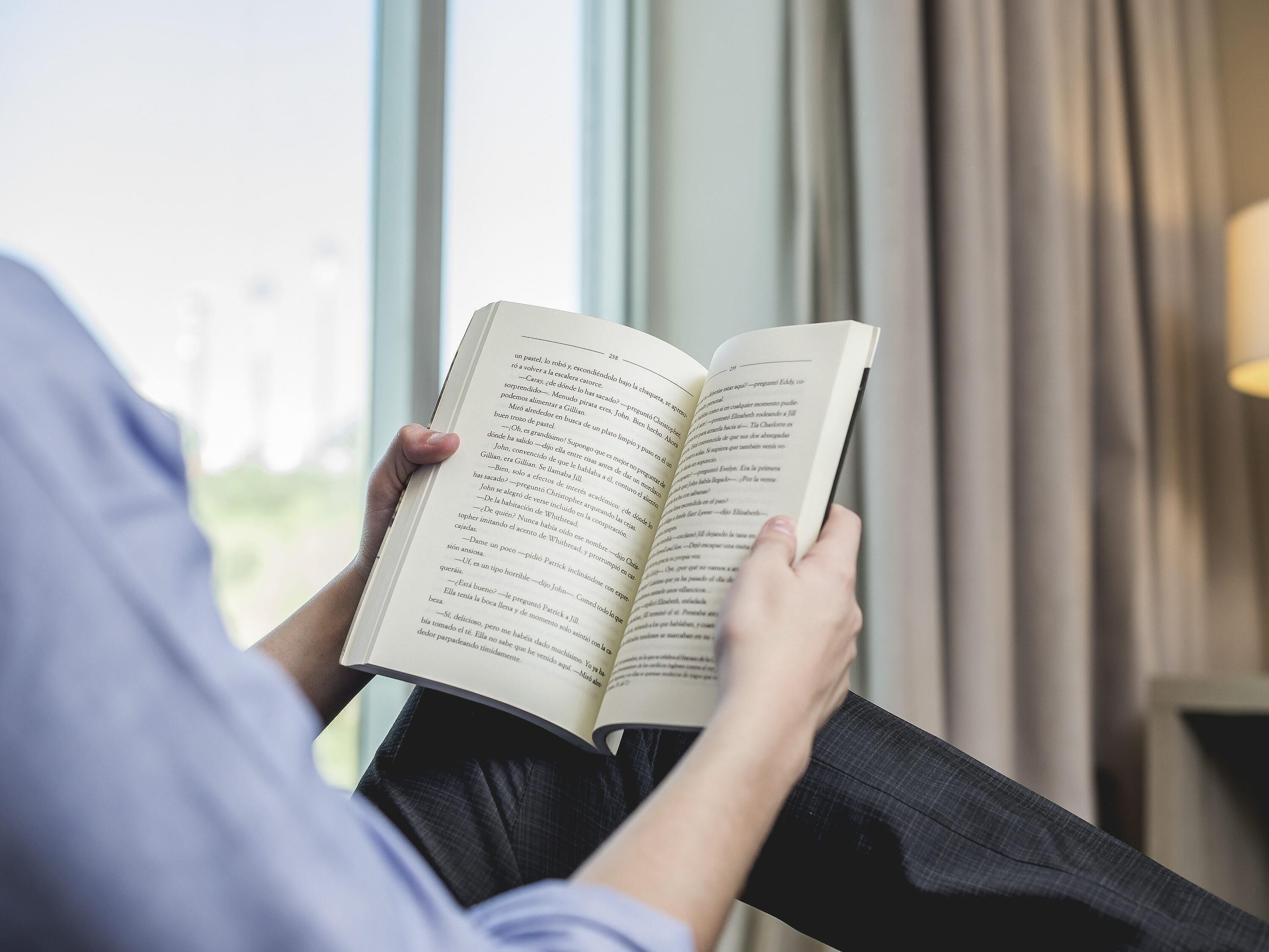 A person reading a book by a window with curtains.
