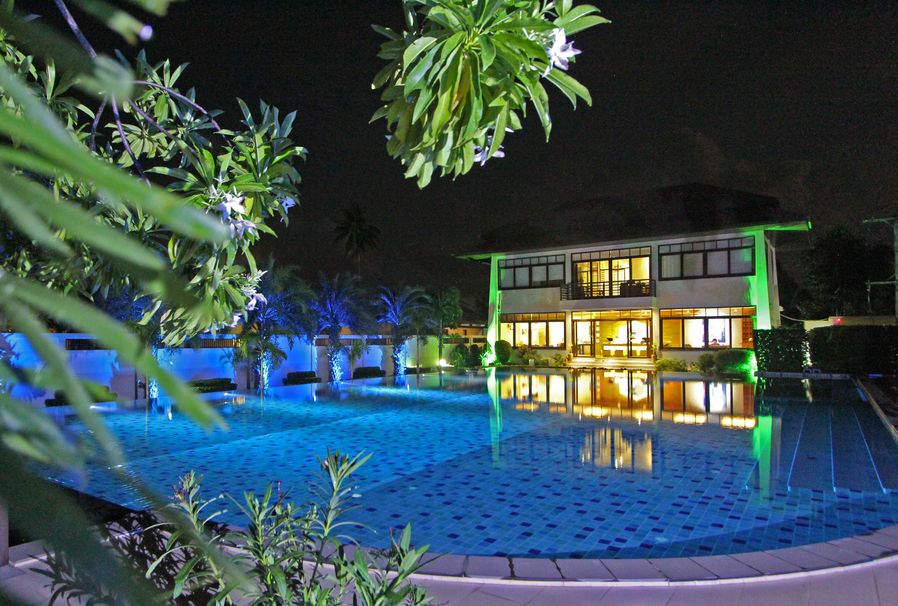 A nighttime view of a hotel with a swimming pool, illuminated by blue and green lights, surrounded by lush greenery.