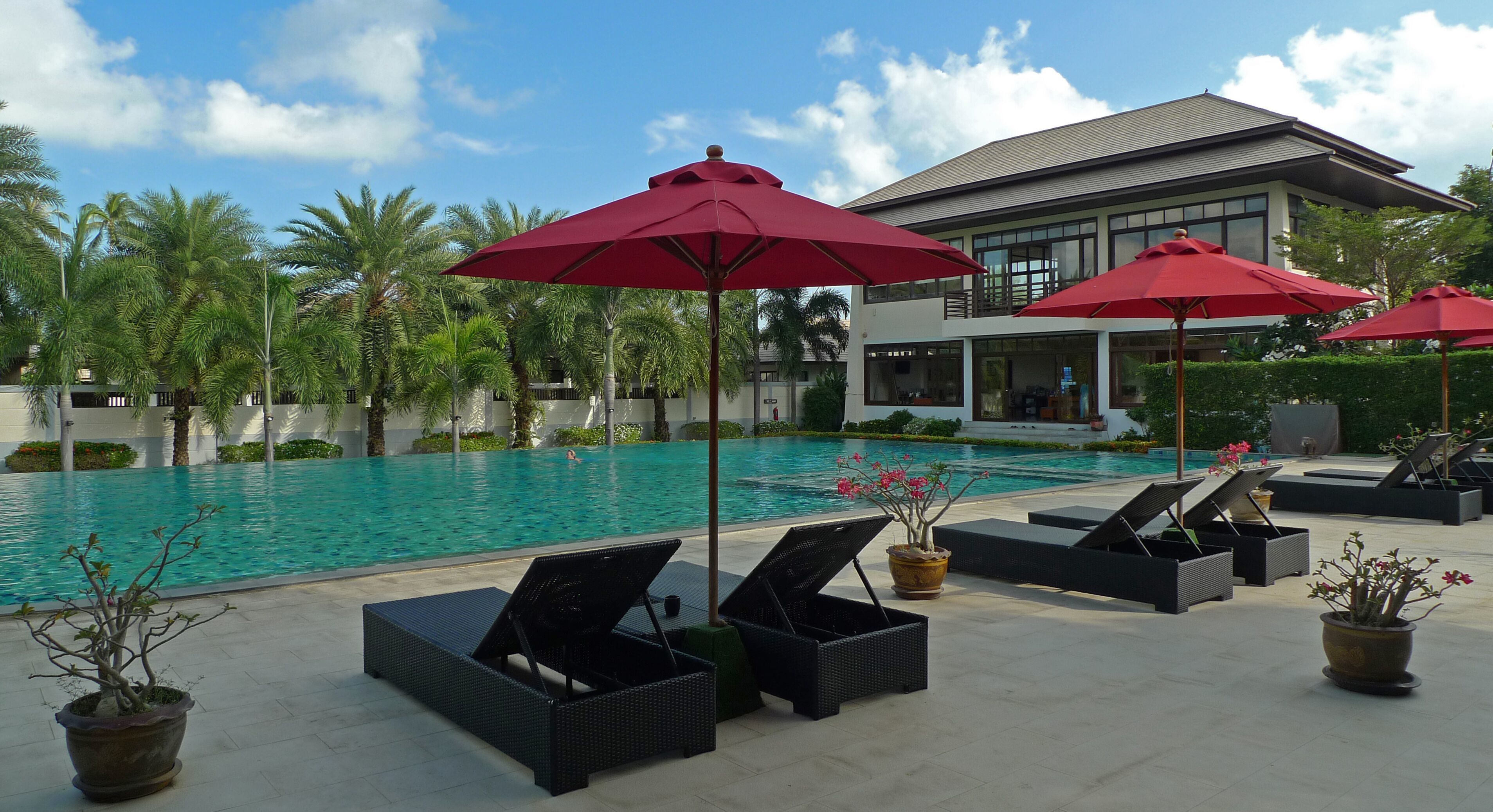 Un espace aménagé au bord de la piscine, avec des chaises longues et des parasols rouges, une piscine à l’eau d’un bleu limpide et, au fond, un bâtiment moderne.
