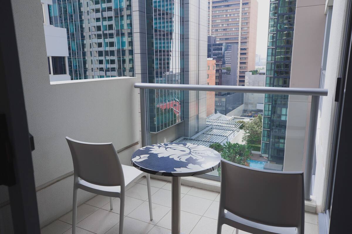 Un balcon avec deux chaises et une table, donnant sur un paysage urbain composé d’immeubles modernes et d’un véhicule rouge.