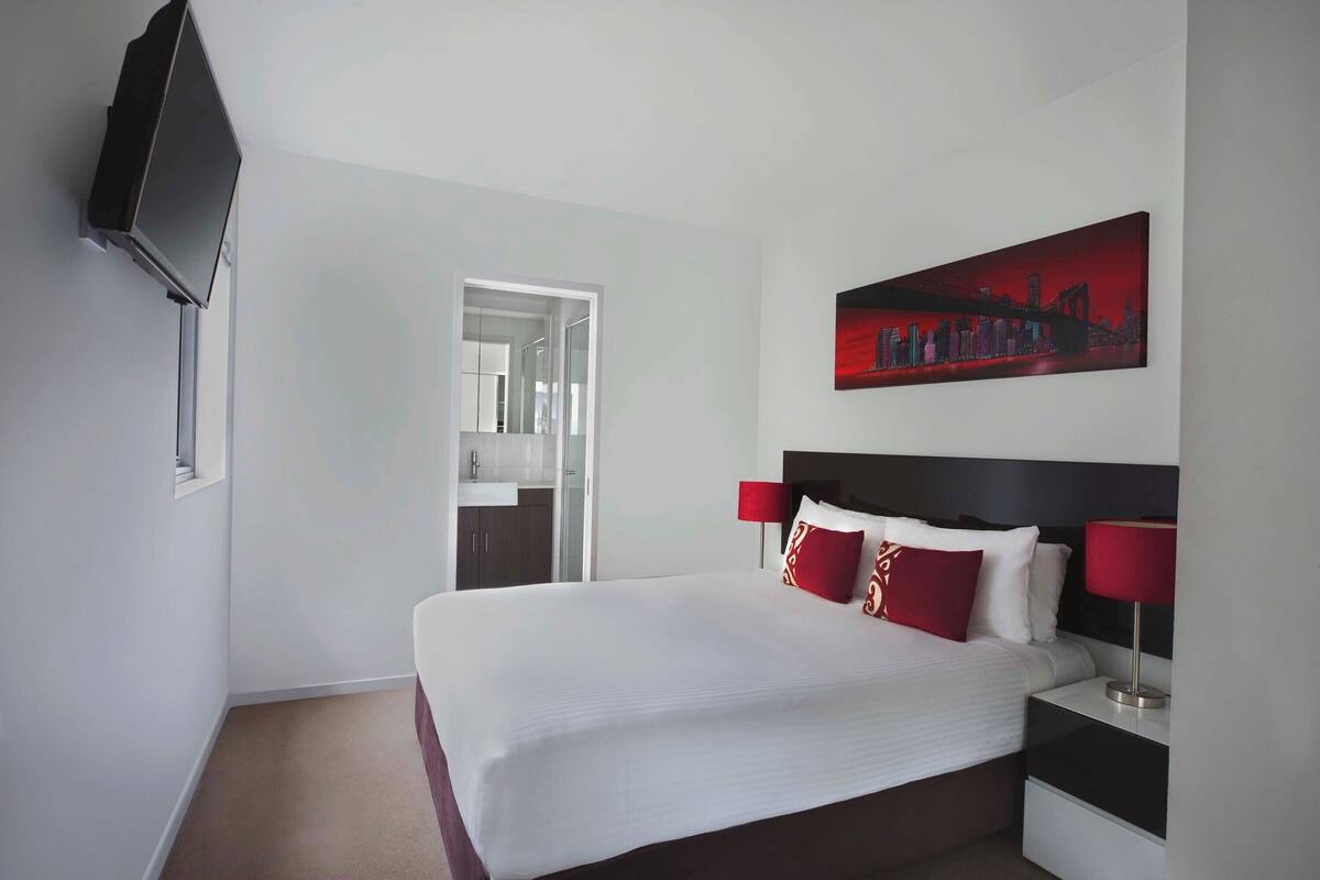 Une chambre d’hôtel avec un grand lit, un téléviseur fixé au mur, un tableau représentant un paysage urbain et une lampe rouge sur la table de chevet.