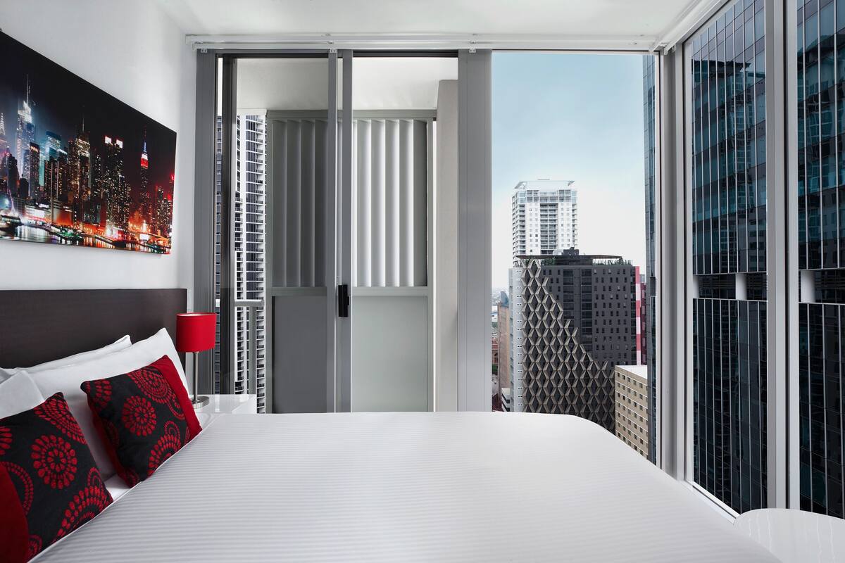 Une chambre d’hôtel avec un lit, une lampe rouge, une vue sur la ville et une grande fenêtre.