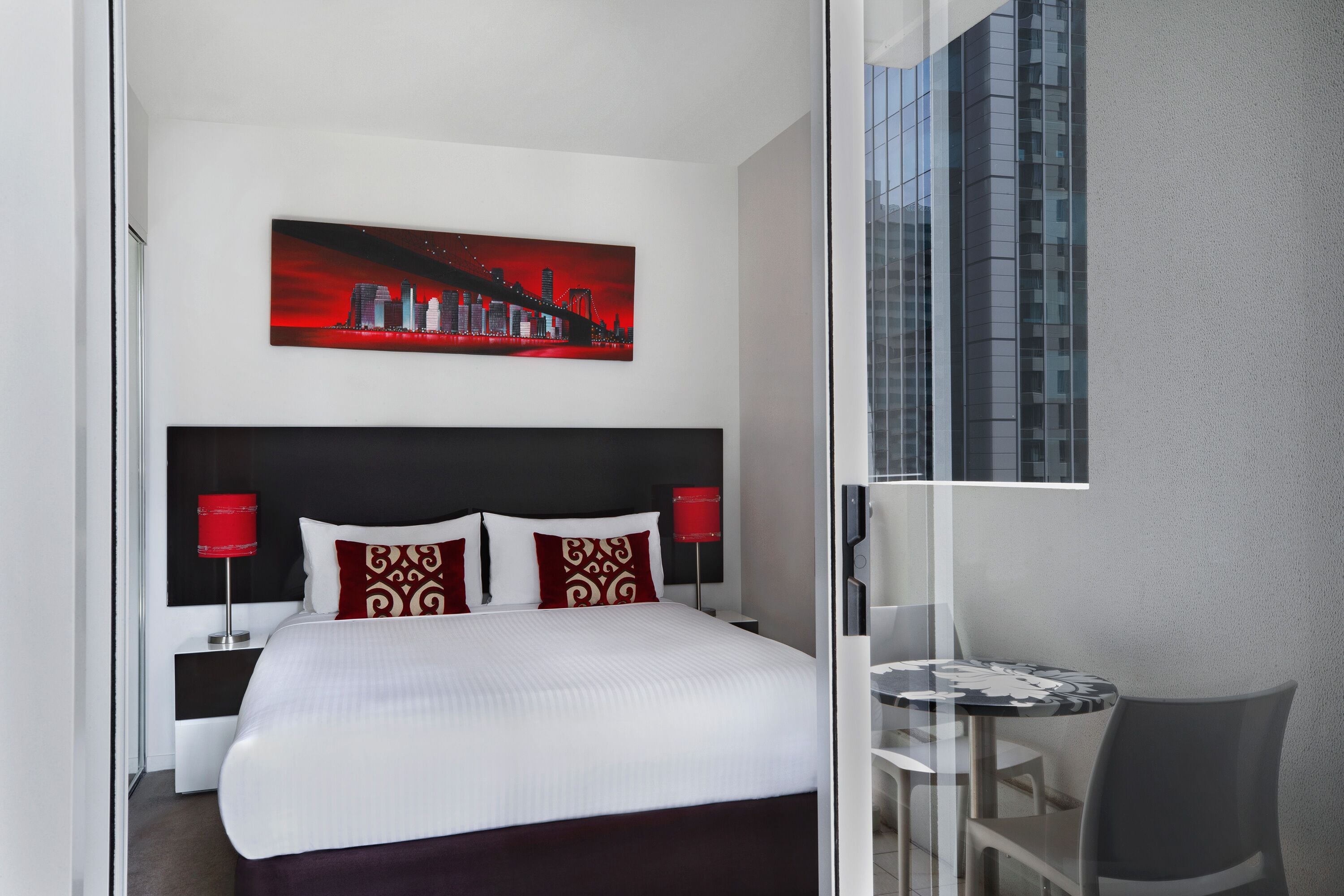 A modern hotel room with a large bed, a cityscape painting, a small round table, and a chair.