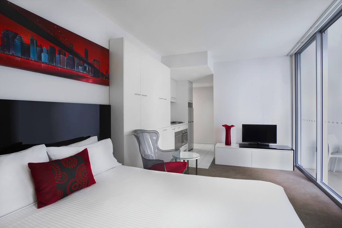 Une chambre d’hôtel moderne avec un grand lit, un bureau, une chaise, un téléviseur et un tableau représentant un paysage urbain accroché au mur.