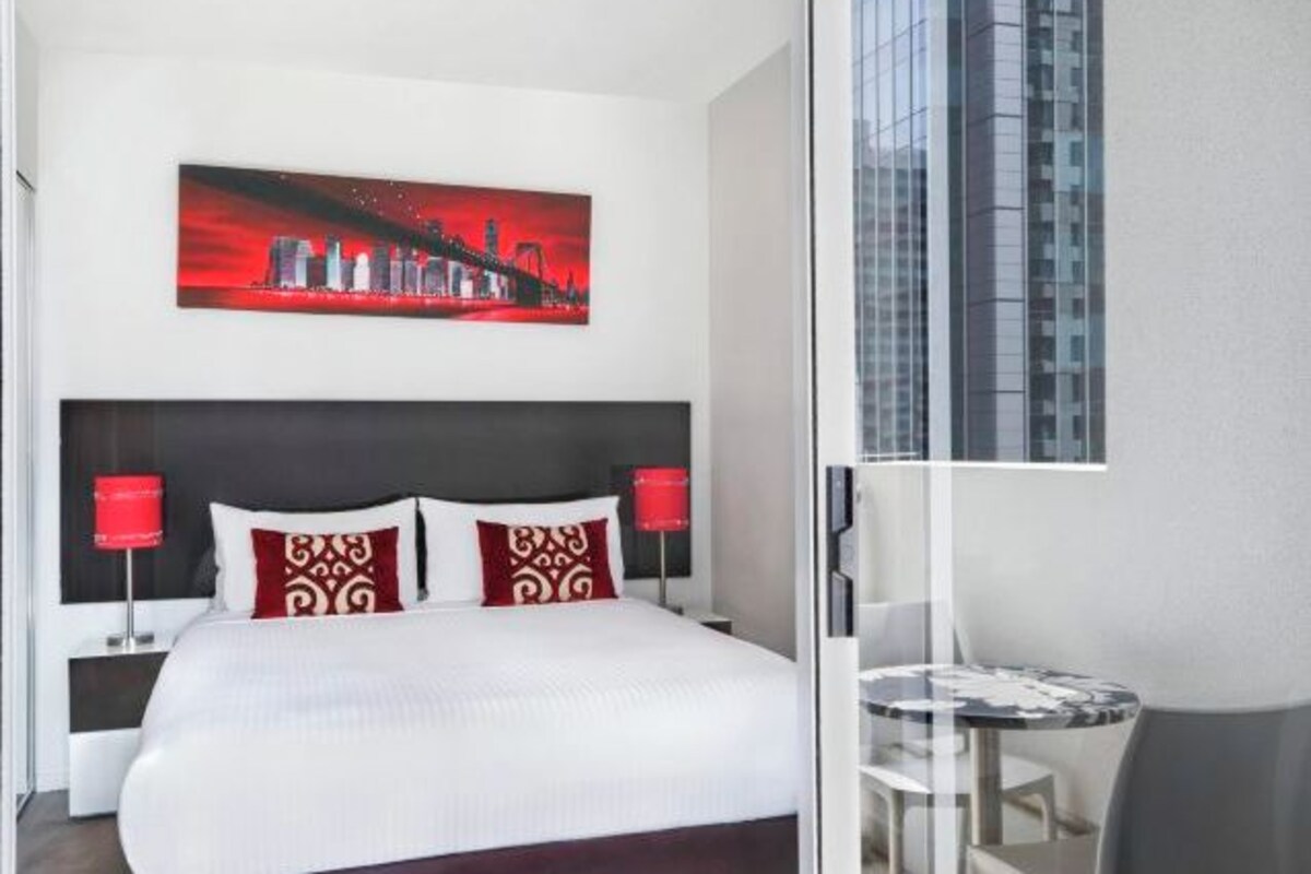 A hotel room with a bed, a red and white cityscape painting, a glass table, and a view of a tall building.