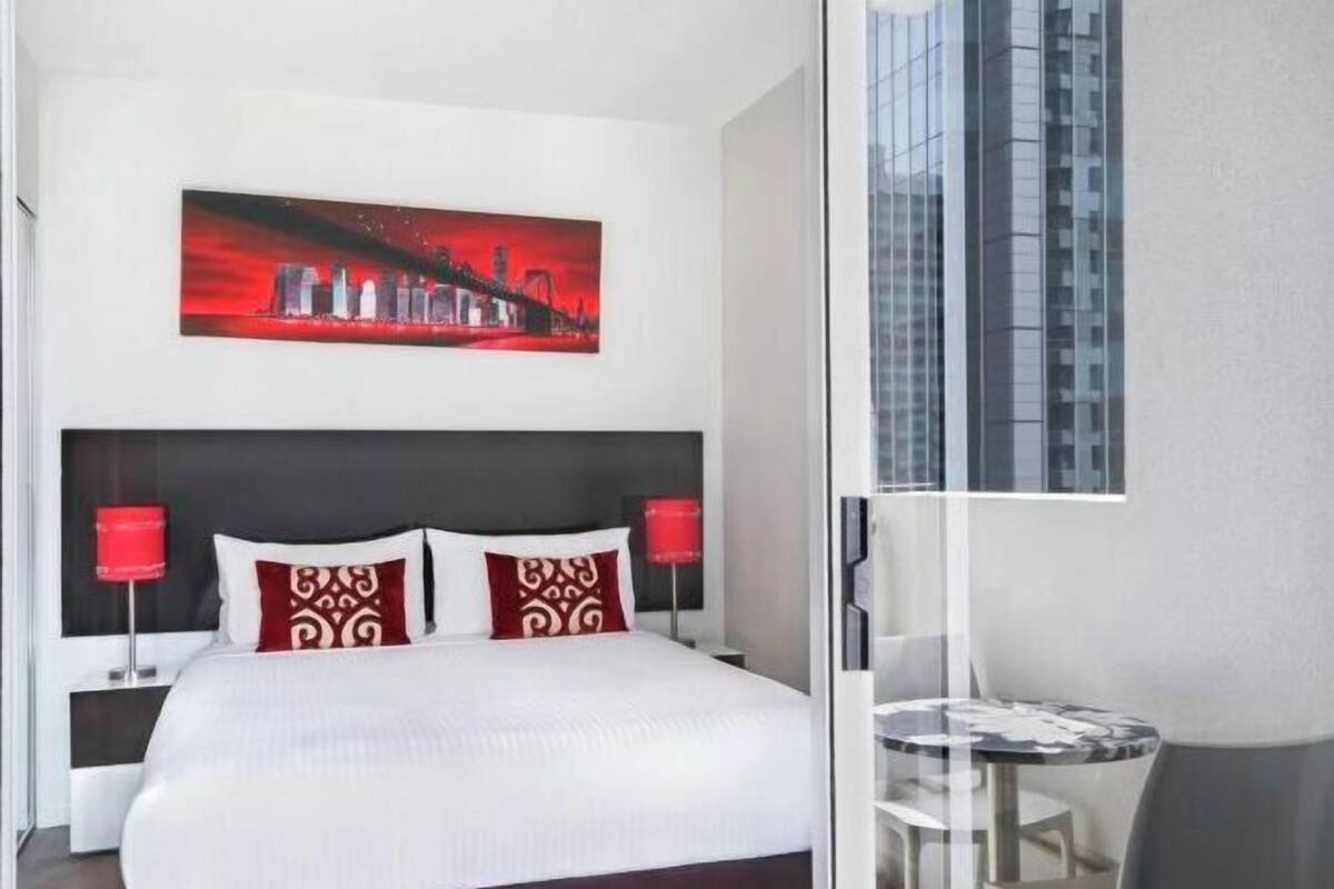 A hotel room with a bed, a red and white cityscape painting, a glass table, and a view of a tall building.