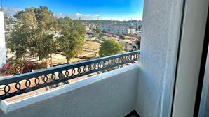 Un balcon doté d’une balustrade en métal, un ciel d’un bleu limpide et une vue sur des bâtiments et des arbres au loin.