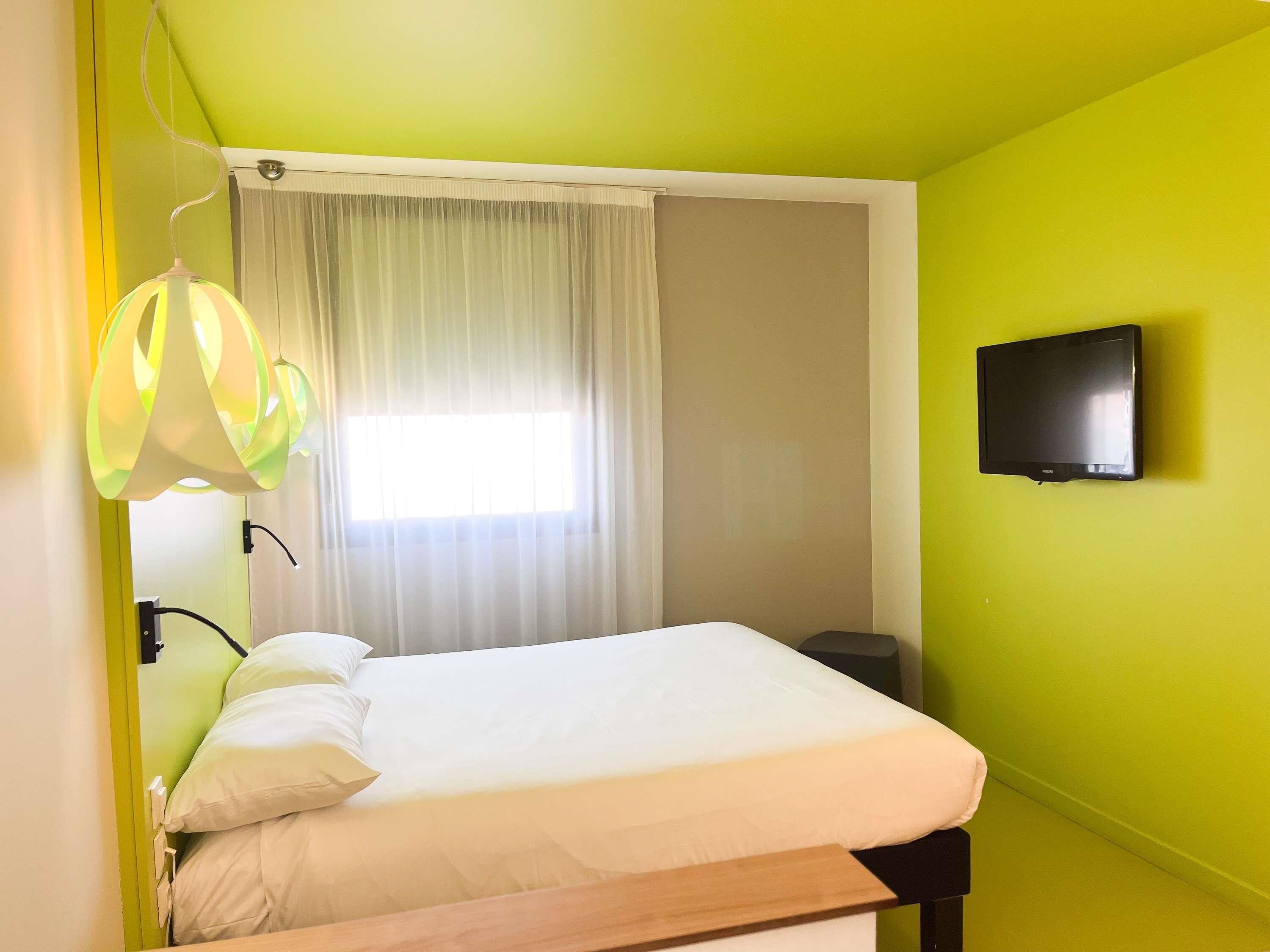 A room with a bed, a television on the wall, and a window with curtains.