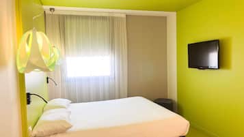 A room with a bed, a television on the wall, and a window with curtains.