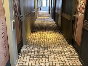 Un pasillo con piso de adoquines, puertas numeradas y un extintor.