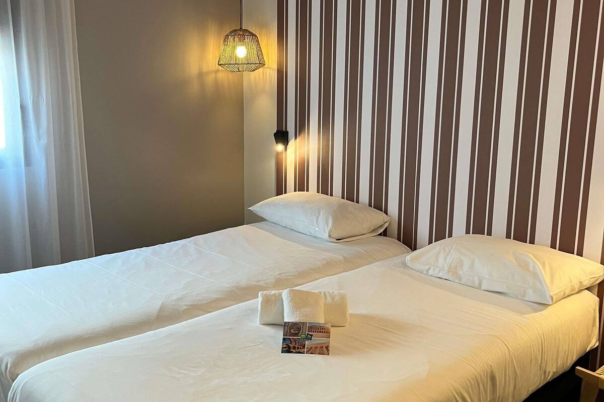 A hotel room with two beds, striped wallpaper, a hanging light fixture, and a small table with a magazine.