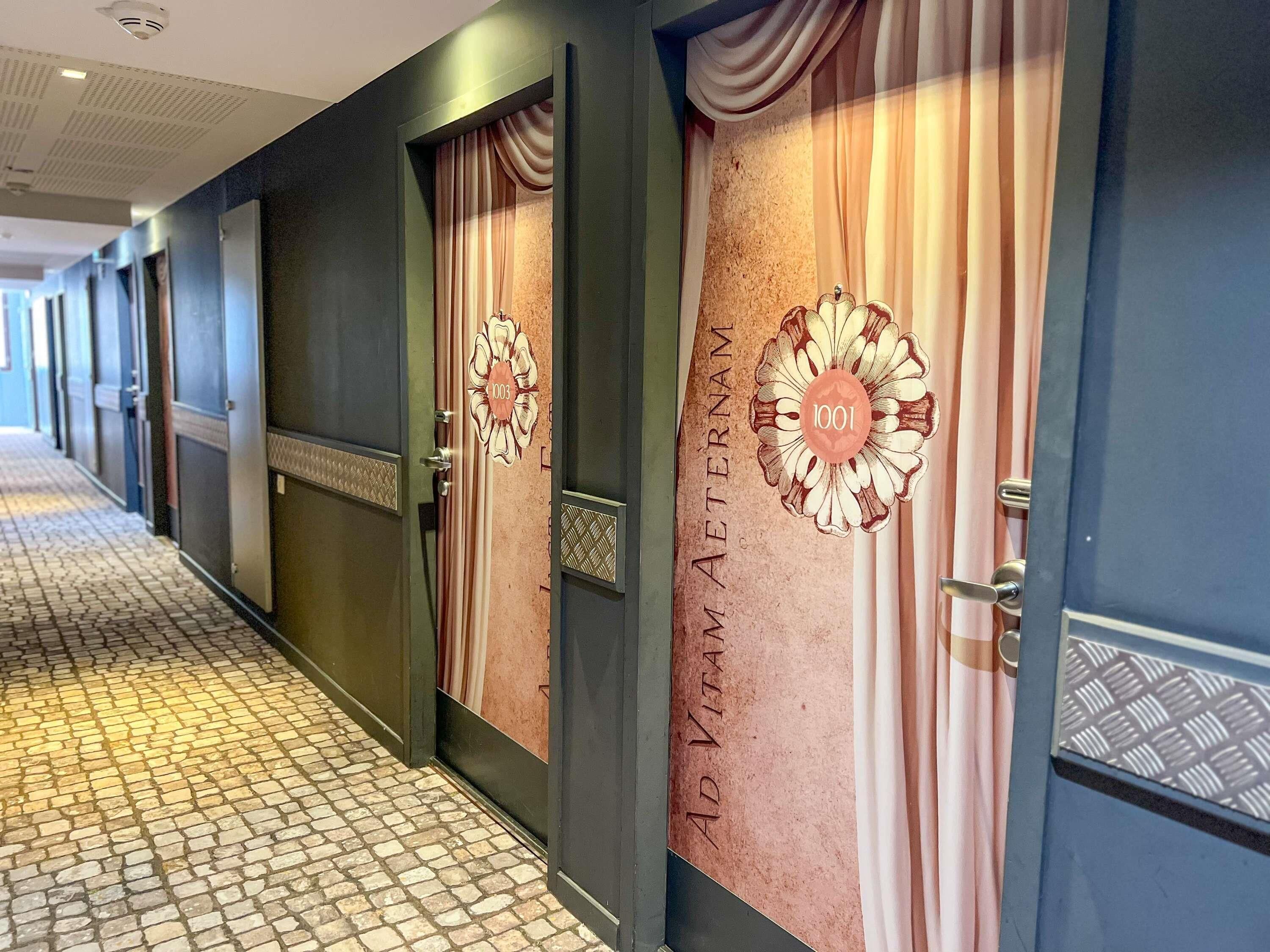 Un pasillo de hotel con puertas que indican los números '1003' y '1001', y un letrero que dice 'AD VITAM METAM'.