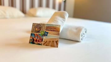 A neatly made bed with a rolled white towel and a small stack of postcards featuring various landmarks.
