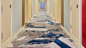Un couloir d’hôtel avec un tapis à motifs, des portes des deux côtés et une porte rouge au bout.