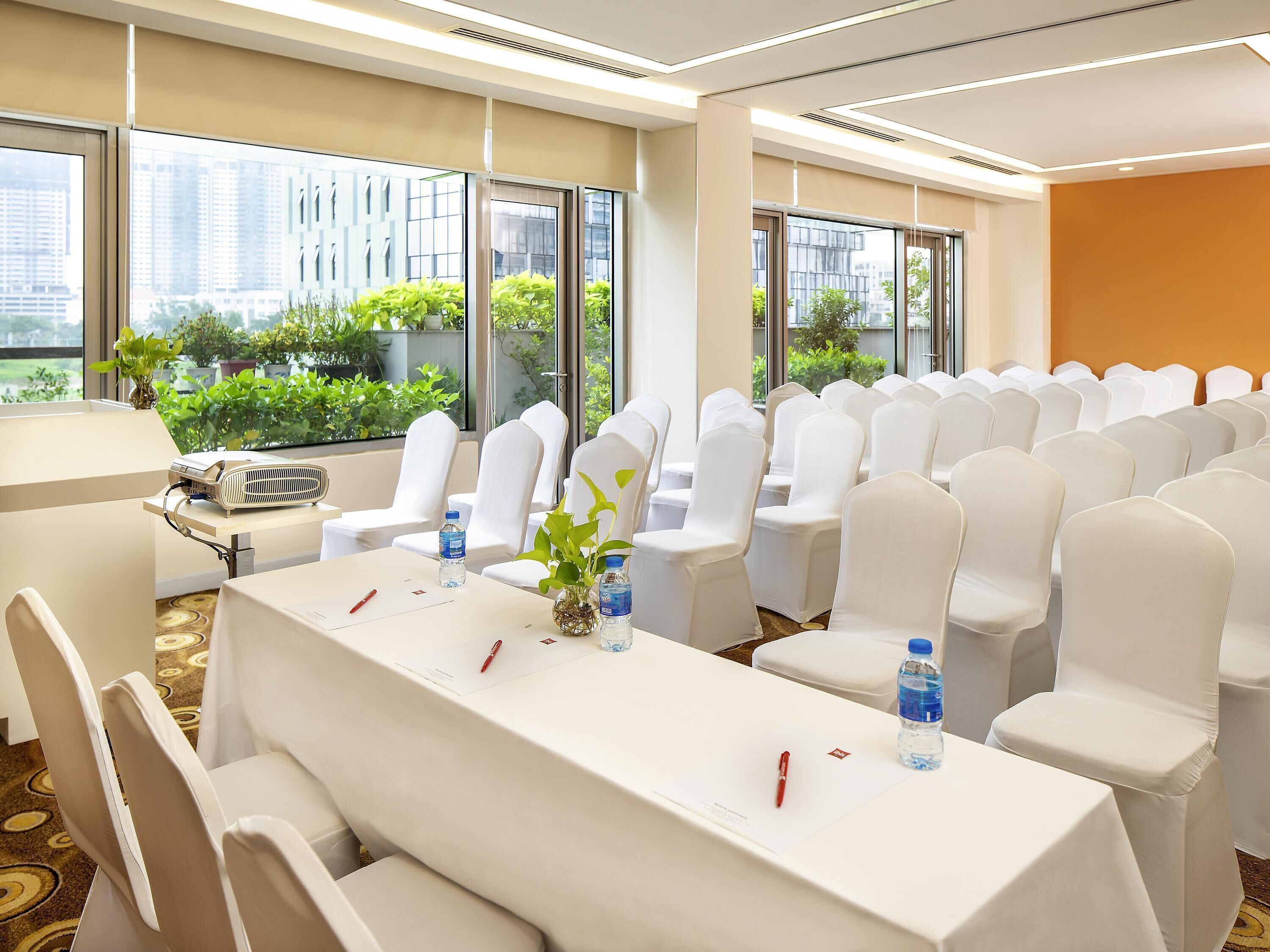 A conference room with white chairs, a long table, and large windows with a view of buildings and greenery.
