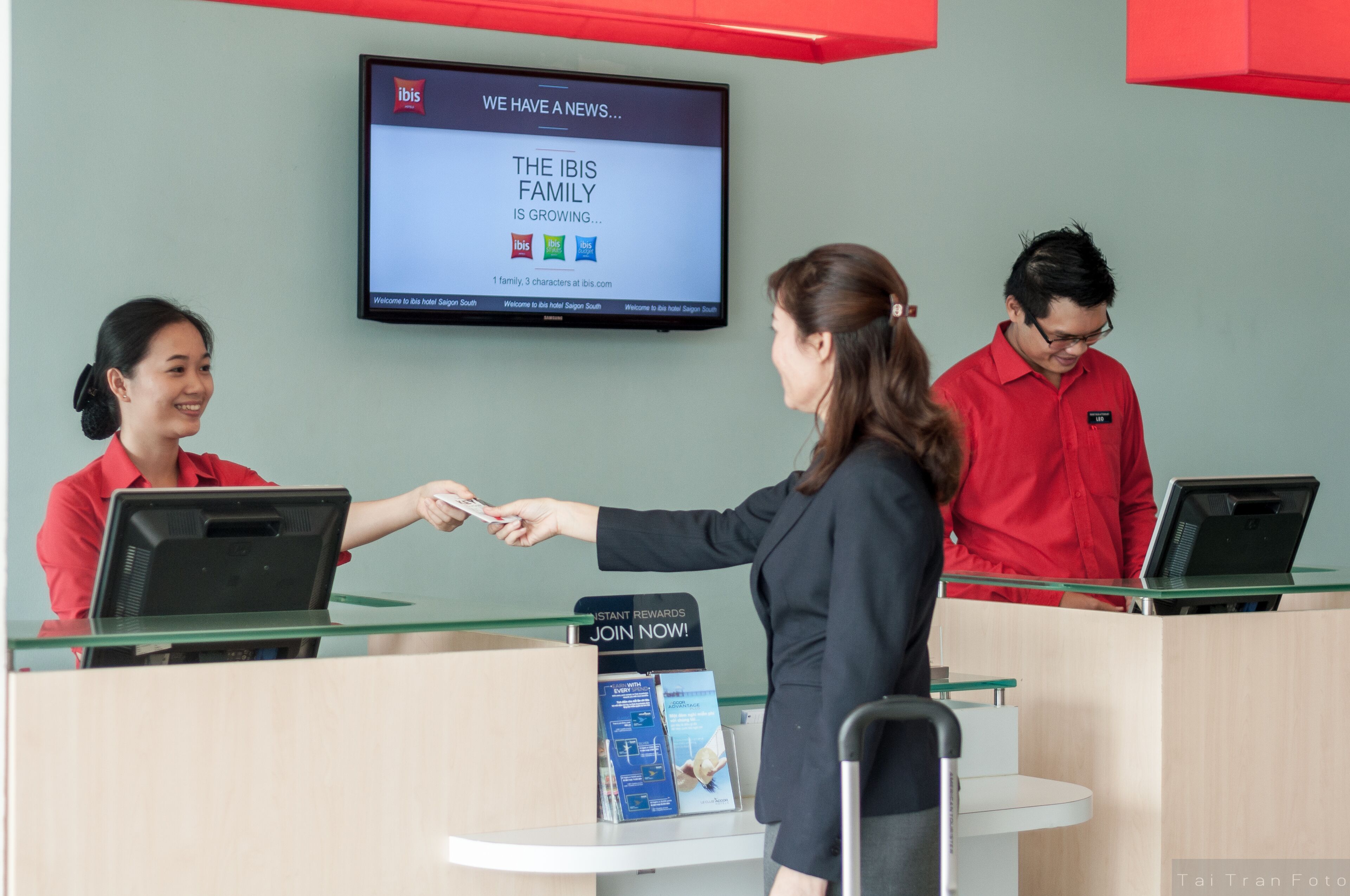 A hotel reception area with a digital screen displaying a news message, a receptionist, and a guest interacting.