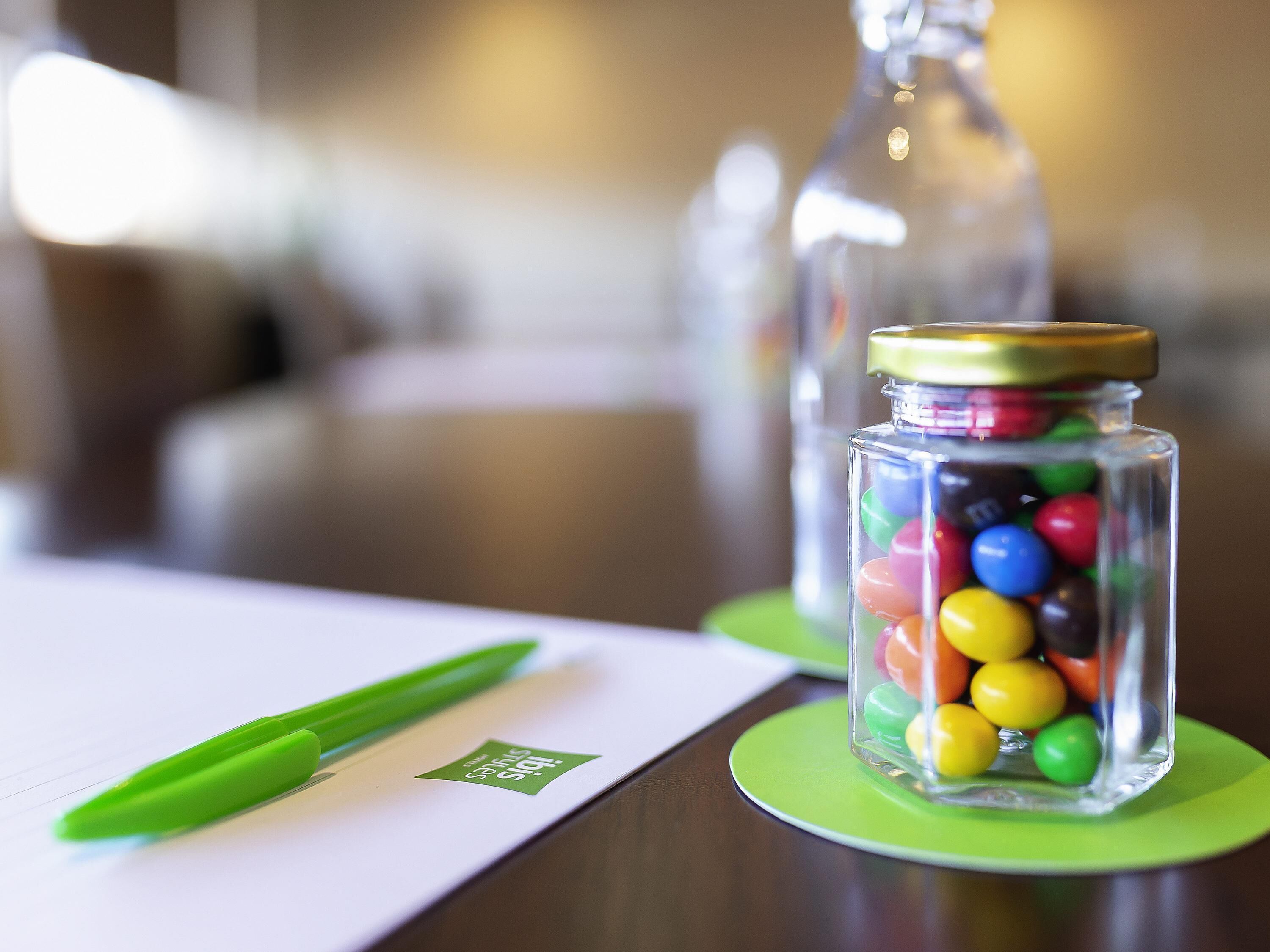 Un frasco de vidrio transparente con caramelos de colores sobre un posavasos verde, un bolígrafo verde sobre una superficie blanca y un fondo desenfocado.