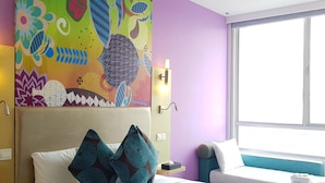 Una habitación de hotel con cama, almohadas, una mesita de noche, una obra de arte en la pared, una ventana y otra mesita auxiliar.