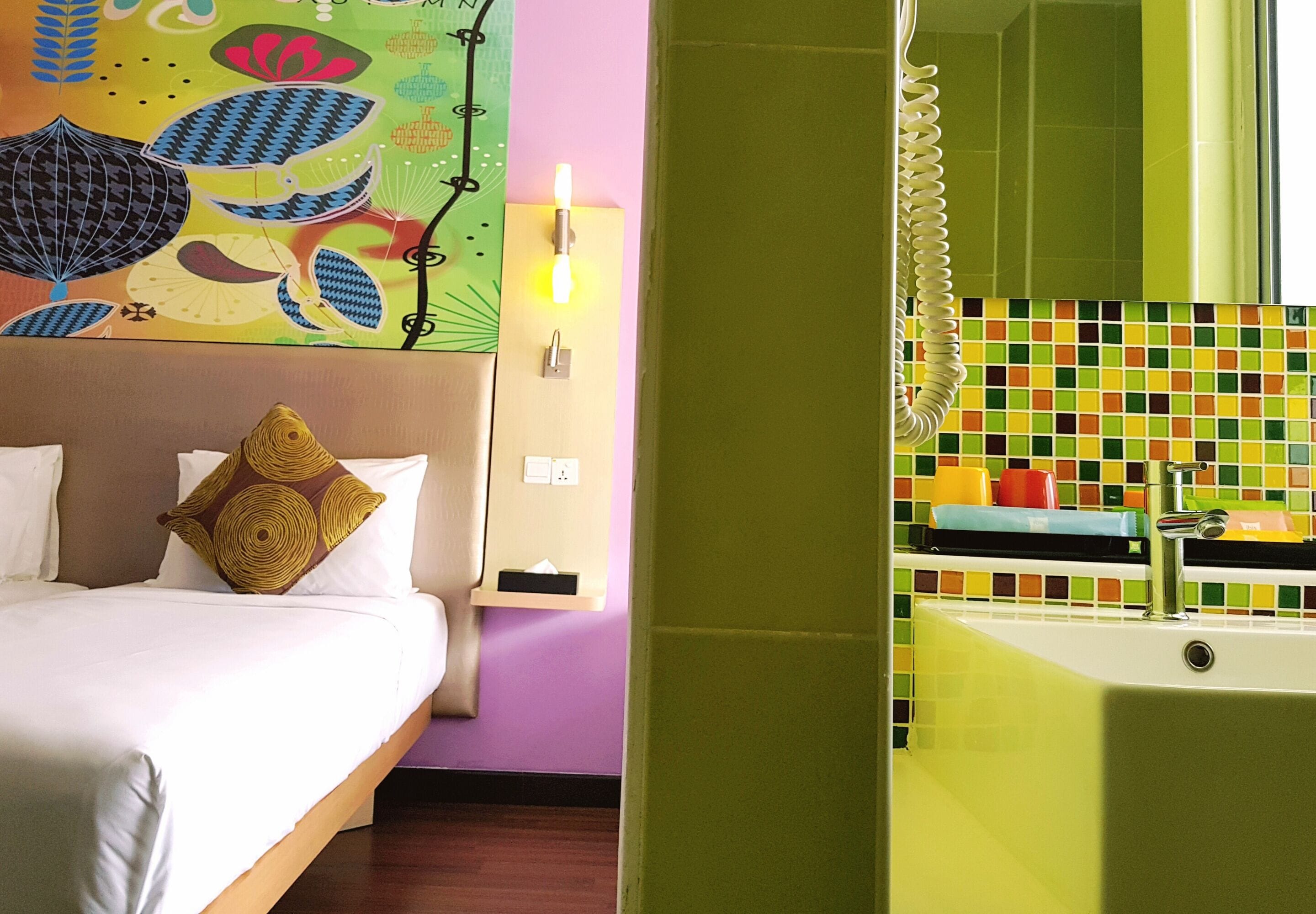 Une chambre d’hôtel comprenant un lit, une table de chevet, une lampe fixée au mur, un tableau abstrait aux couleurs vives, et une salle de bain avec un mur carrelé et une baignoire.