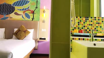 Une chambre d’hôtel comprenant un lit, une table de chevet, une lampe fixée au mur, un tableau abstrait aux couleurs vives, et une salle de bain avec un mur carrelé et une baignoire.