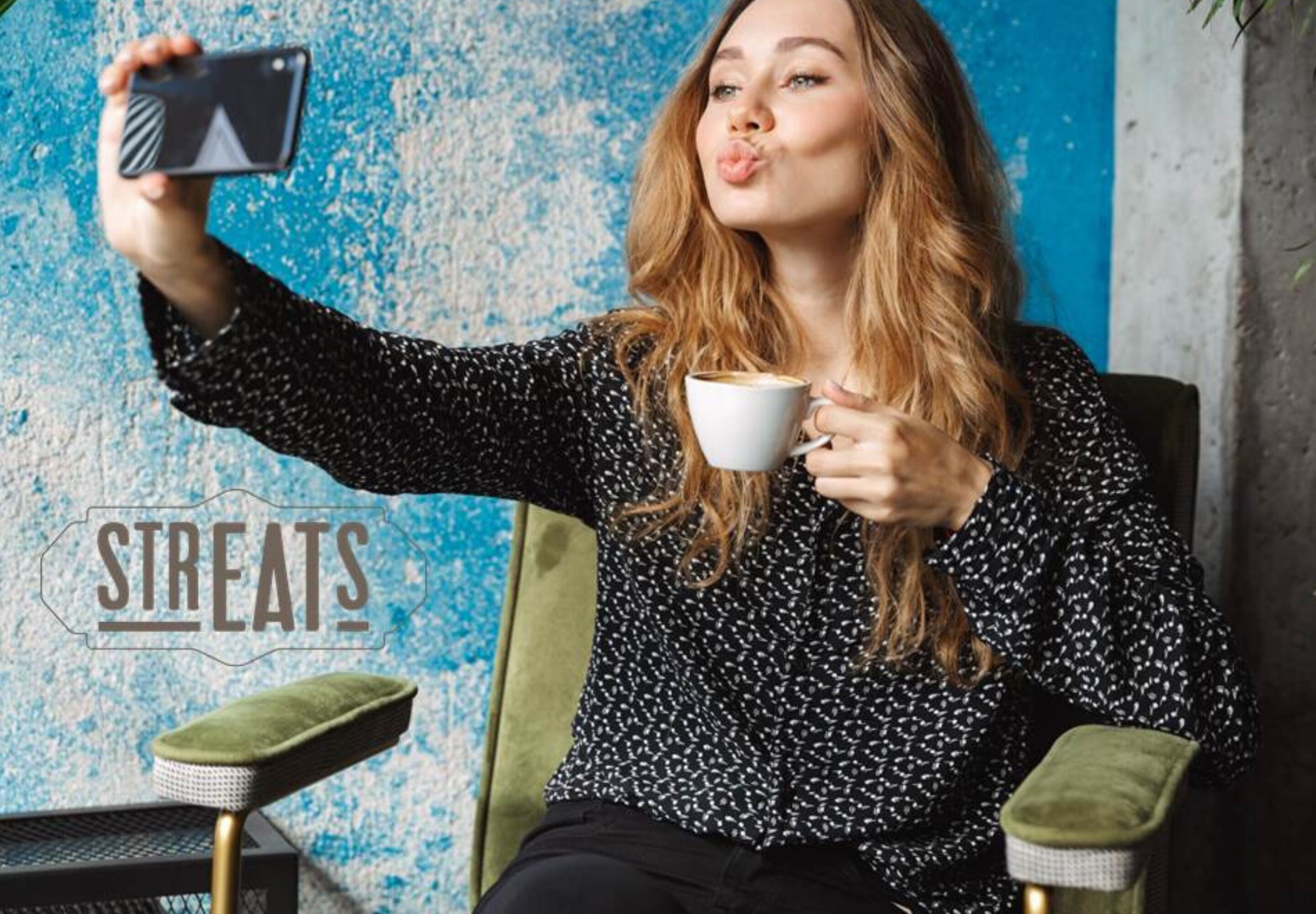 Een persoon zittend in een stoel met een smartphone en een kopje, voor een blauwe wand met een structuur, waar de tekst 'STREATS' op staat.