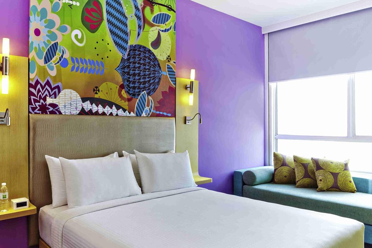 Habitación de hotel con una cama grande, un sofá, un mural de pared colorido y una ventana.