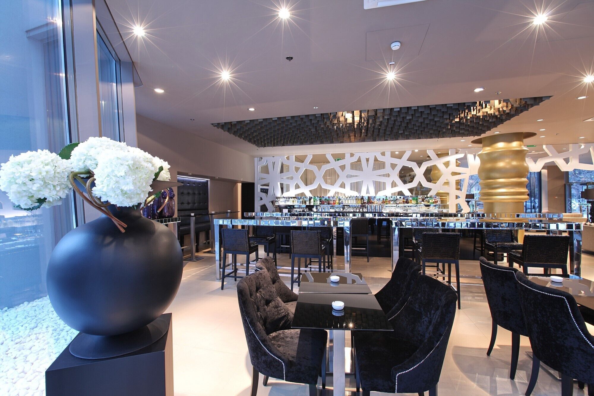 A modern bar area with a high ceiling, black upholstered chairs, and a decorative wall with geometric patterns.