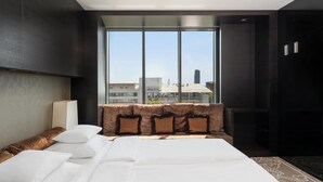 Ein modernes Hotelzimmer mit einem großen Bett, Blick auf die Stadt durch ein großes Fenster und einer gemütlichen Sitzecke mit Kissen.