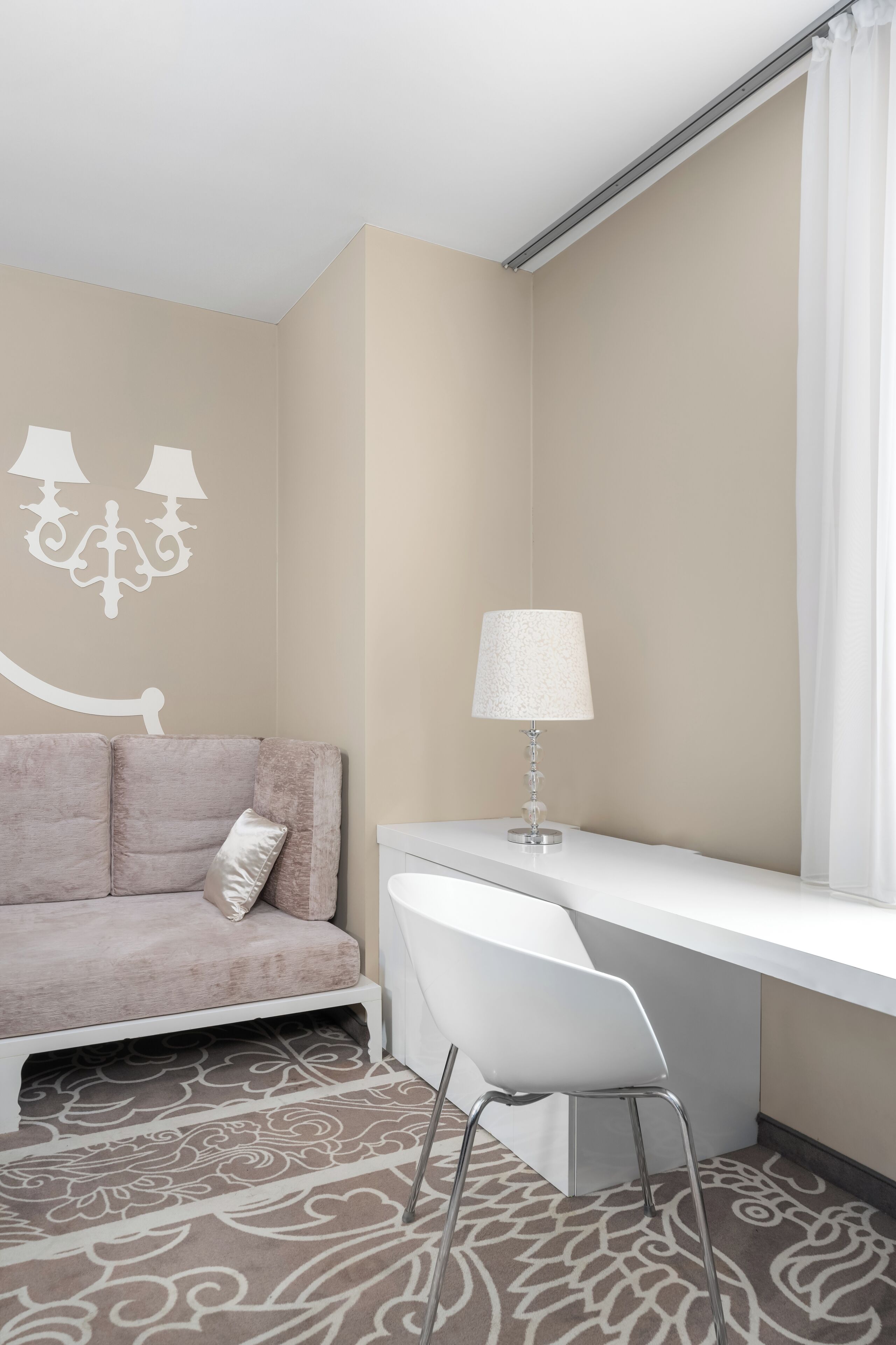 A modern hotel room with a beige wall, a white desk, a white chair, a patterned carpet, and a decorative wall piece.