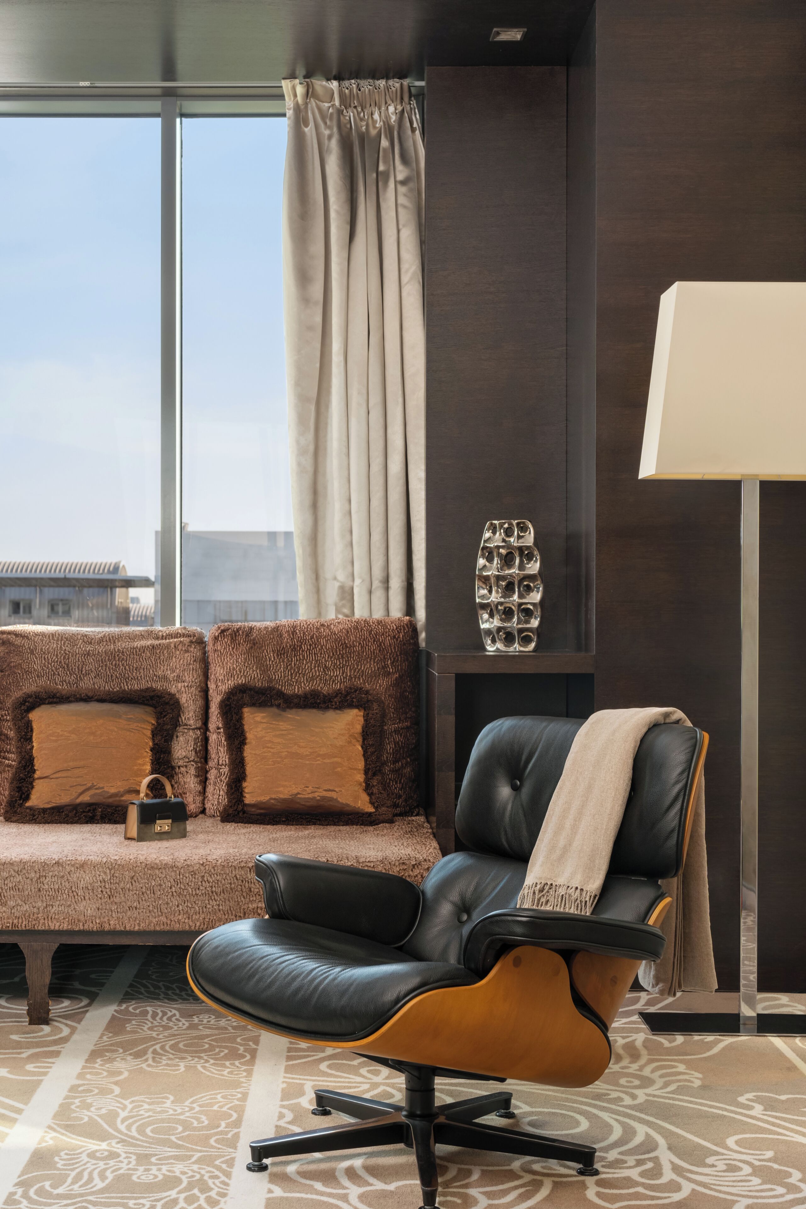 Una sala de estar moderna con un sillón de cuero, un sofá con cojines estampados, un adorno en la pared y una lámpara de pie.