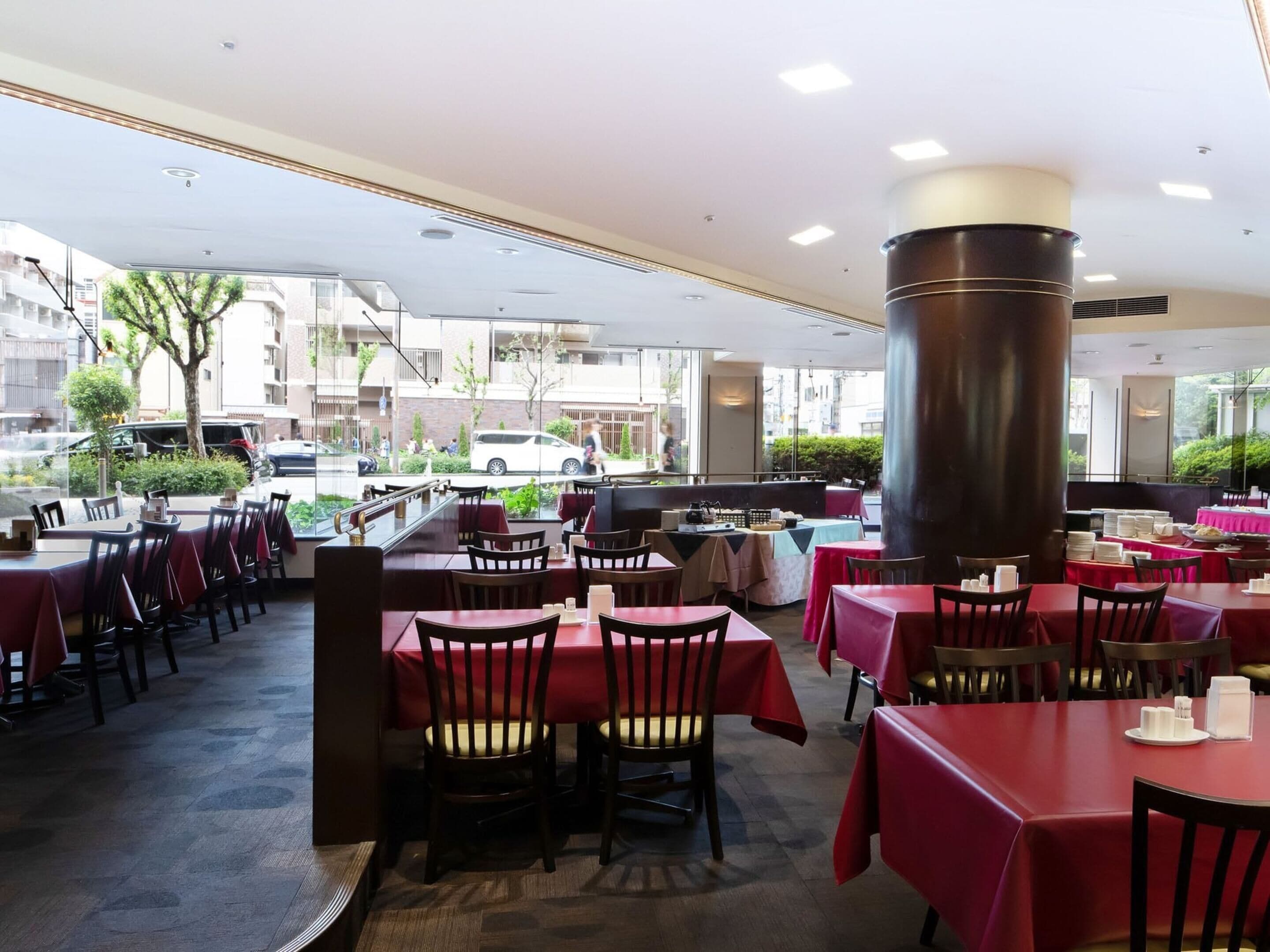 An indoor restaurant with tables covered in red tablecloths and chairs, large windows offering a view of the street outside.
