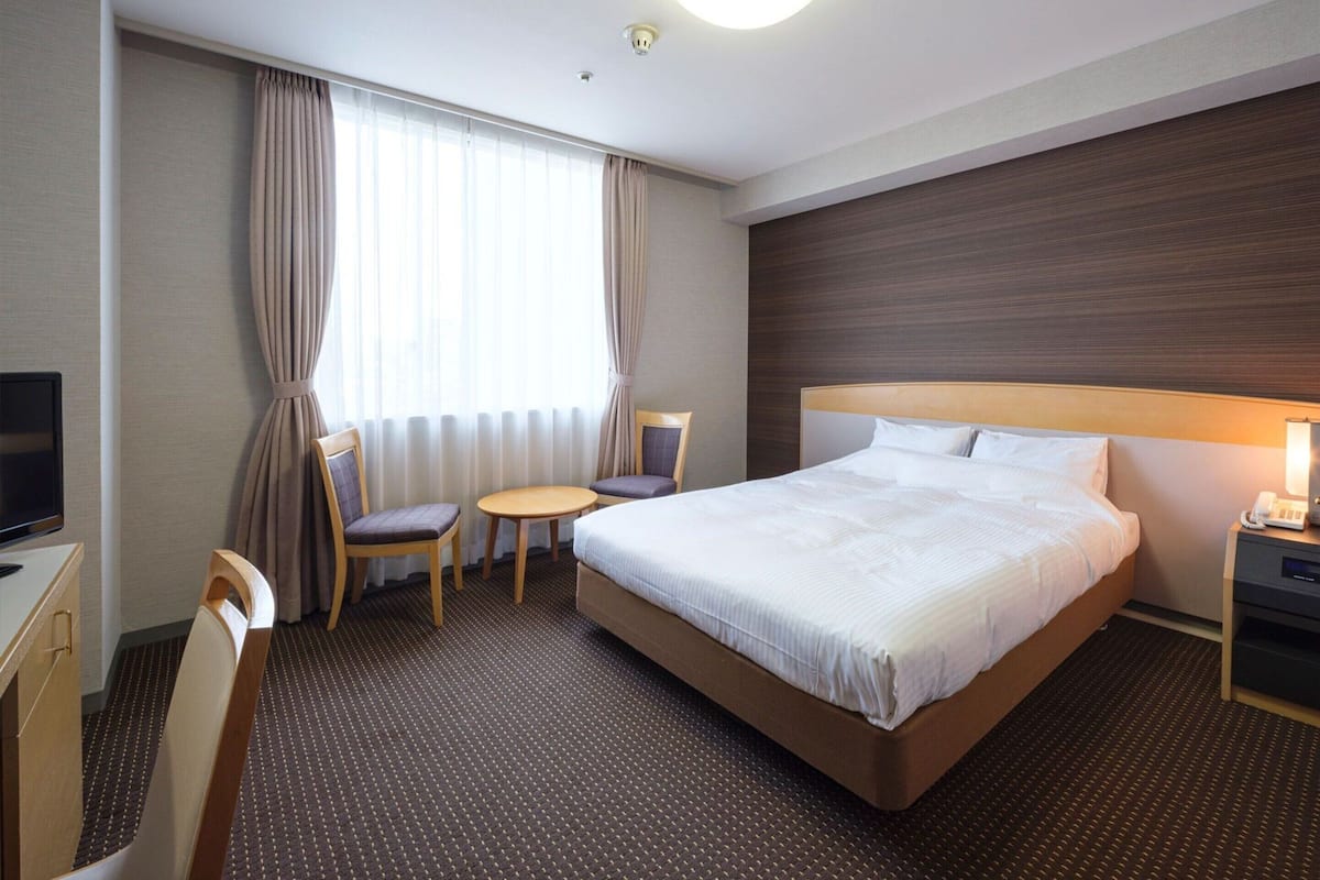 A hotel room with a bed, a desk, a chair, a small table, and a TV.