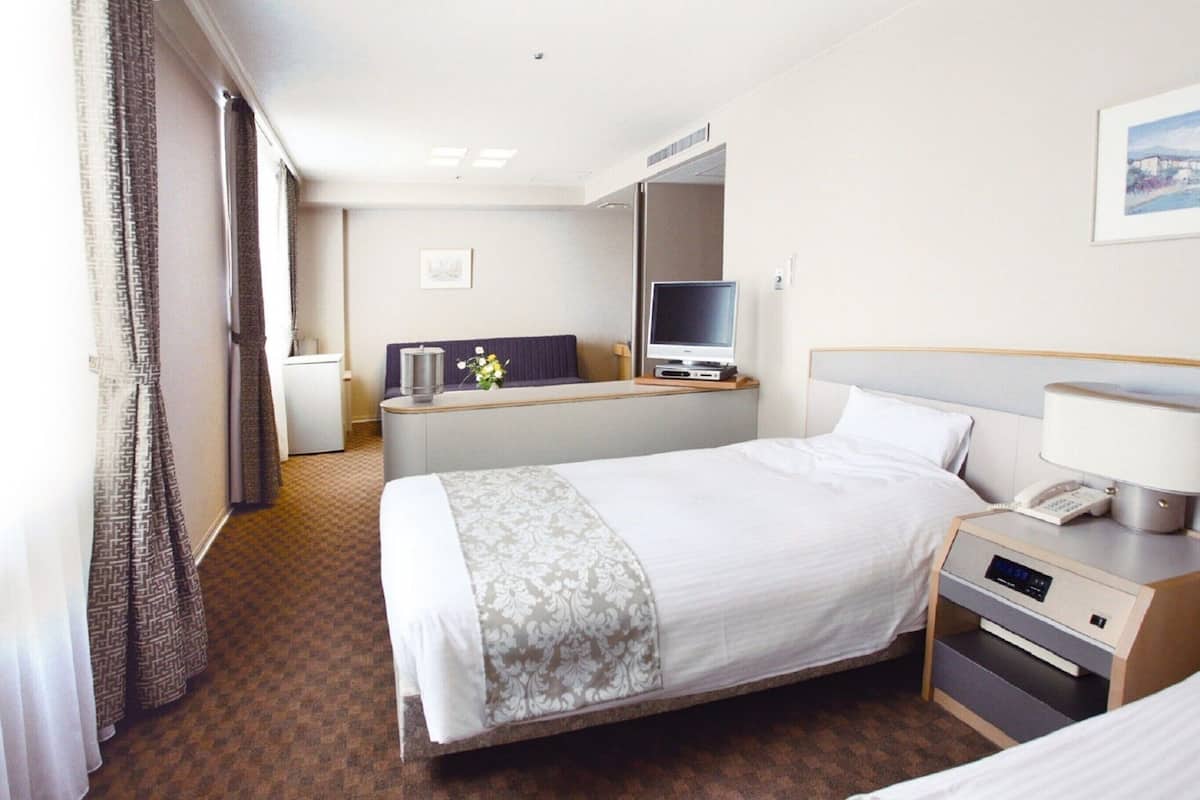 A hotel room with a bed, bedside table, lamp, TV, and a small desk with a chair.