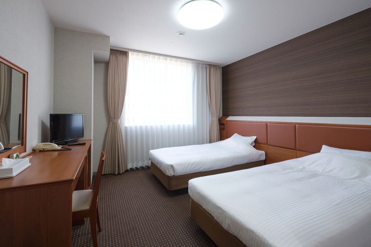 A hotel room with two beds, a desk with a telephone, a television, and a large window with curtains.