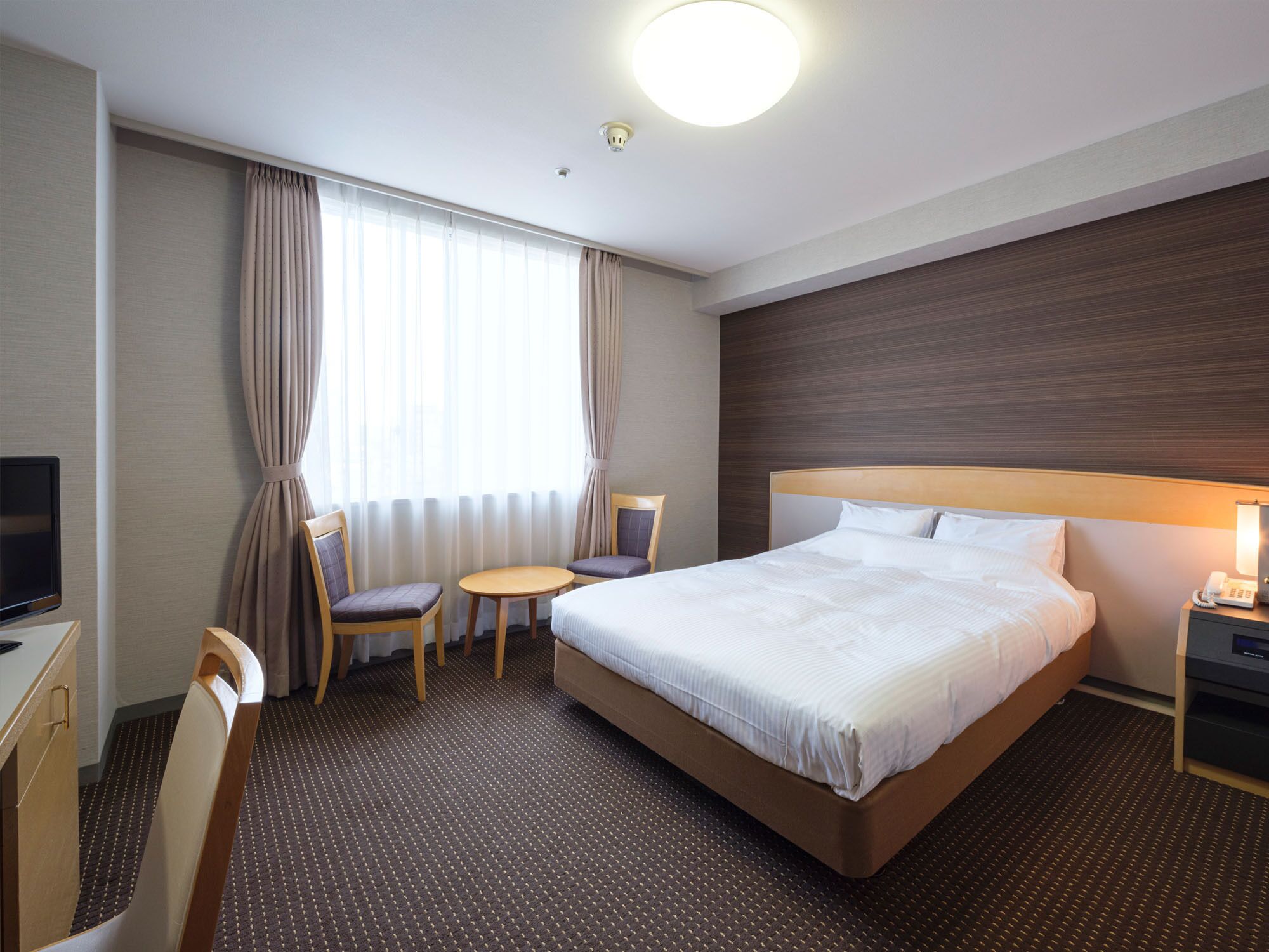 A hotel room with a bed, a desk, a chair, a small table, and a TV.