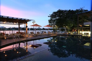 A serene outdoor pool area with lounge chairs and umbrellas, a wooden deck, and a view of the city skyline at dusk. - Riva Surya Bangkok (Bangkok)