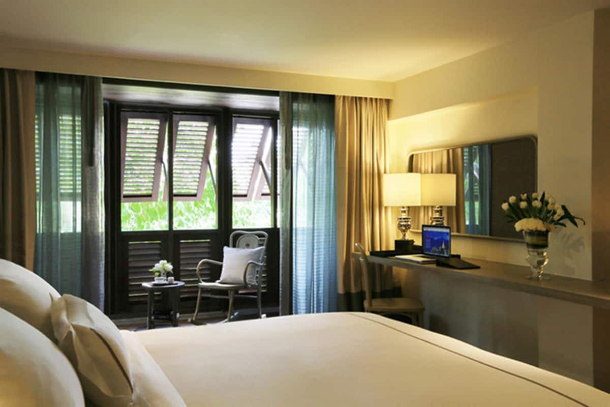 A hotel room with a bed, a desk with a computer, a chair, a small table with a vase of flowers, and a window with curtains.