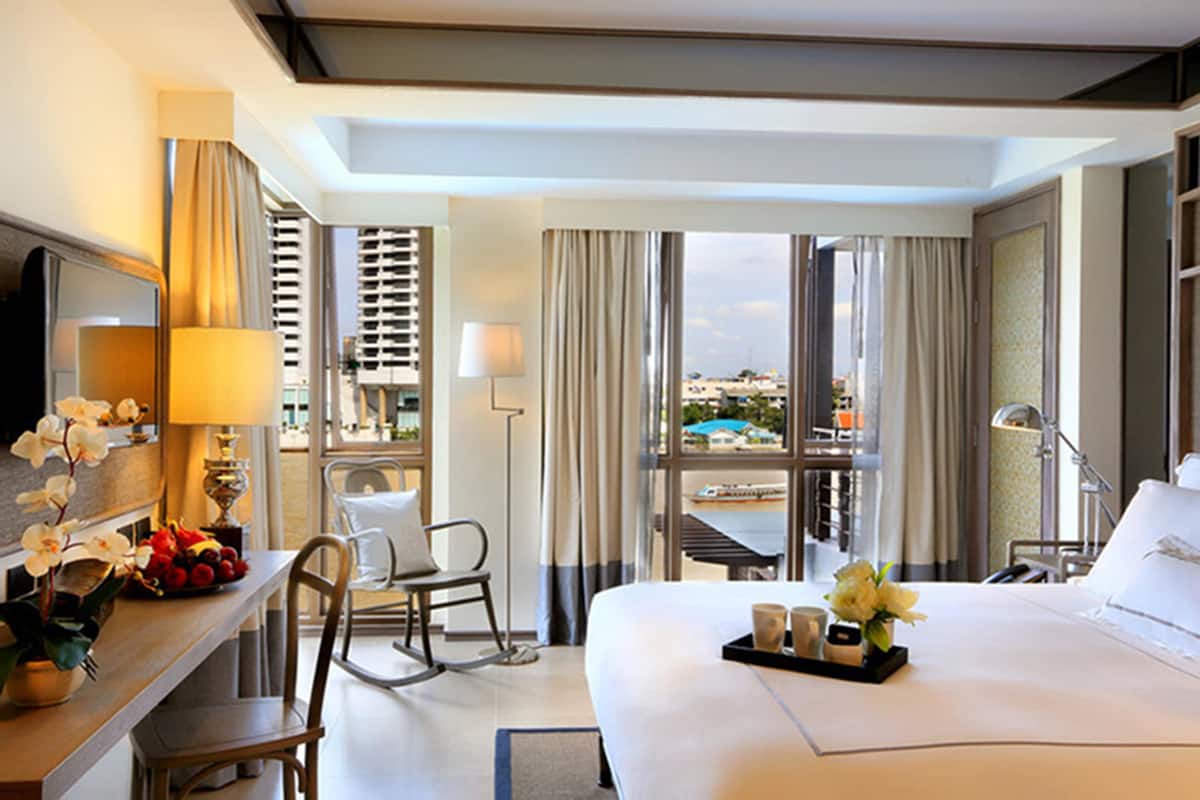 A hotel room with a bed, a desk with a lamp, a chair, a window with curtains, and a view of the outside.