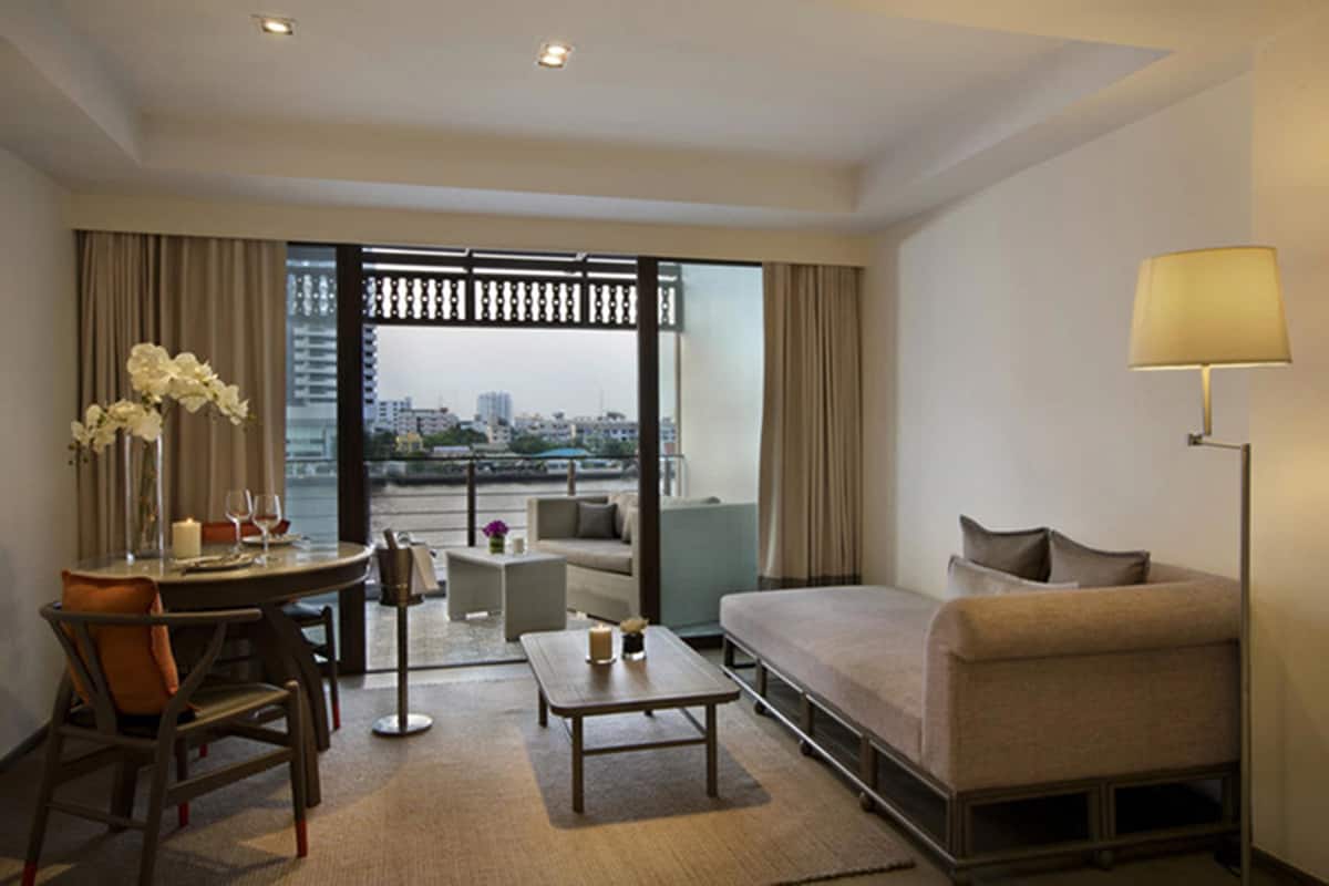 A hotel room with a bed, a sofa, a dining table with chairs, a lamp, and a view of the city through a large window.