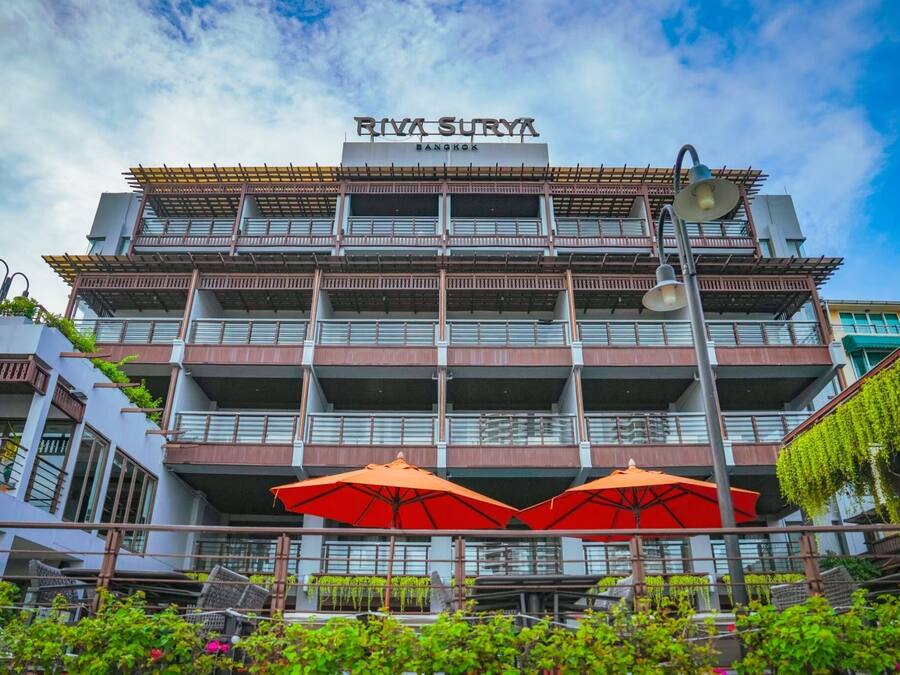 A multi-story building with balconies, a sign reading "RIVA SURYA BANGKOK," and outdoor umbrellas.