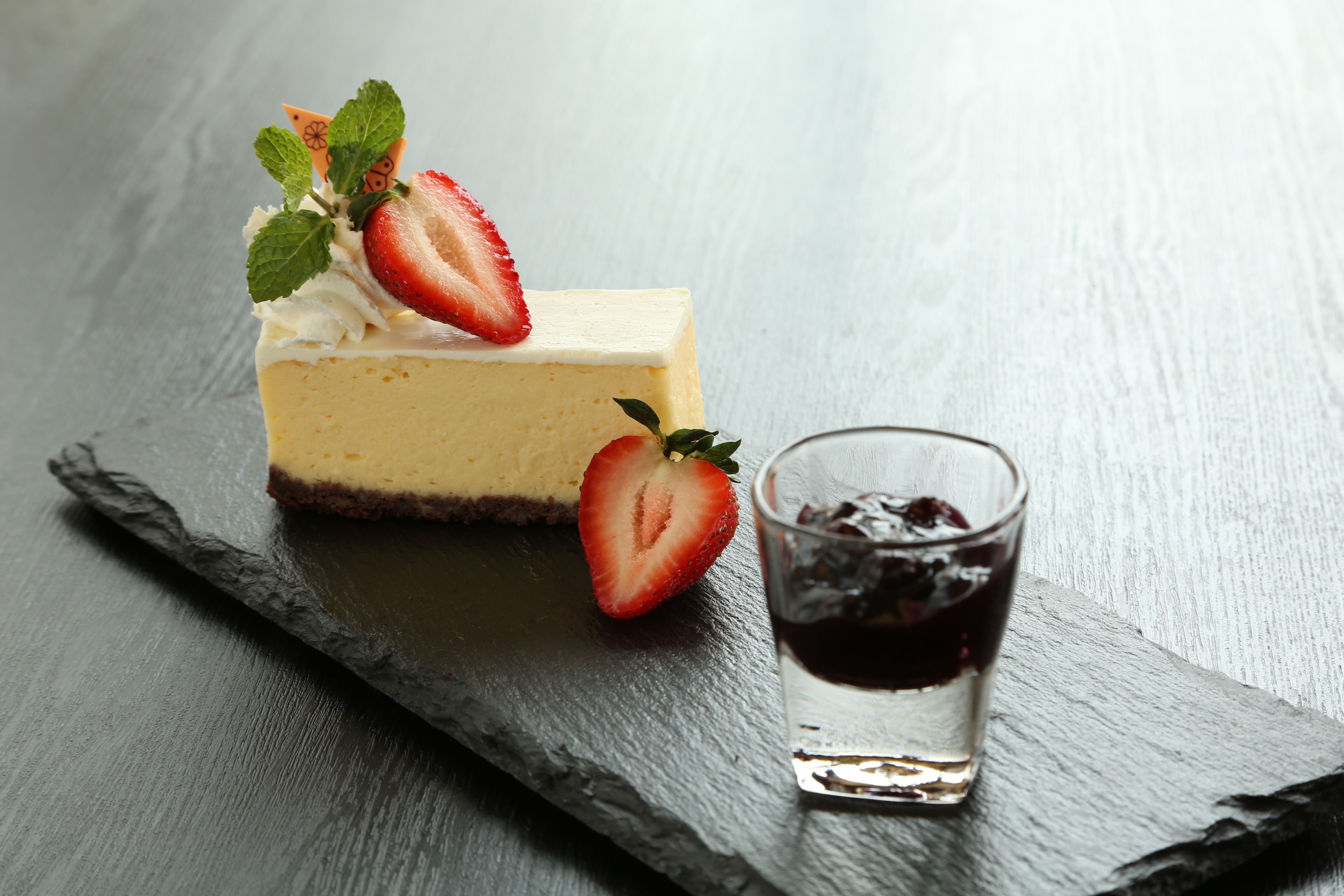 A slice of cheesecake with strawberries and mint on a slate serving board, accompanied by a glass of dark liquid.