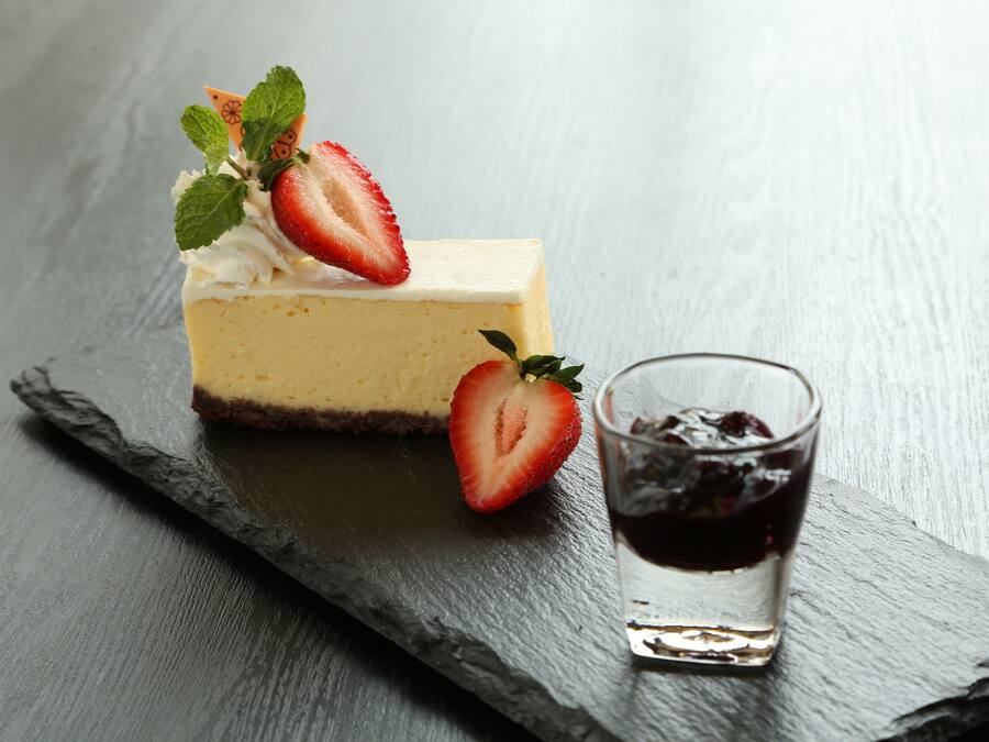 A slice of cheesecake with strawberries and mint on a slate serving board, accompanied by a glass of dark liquid.