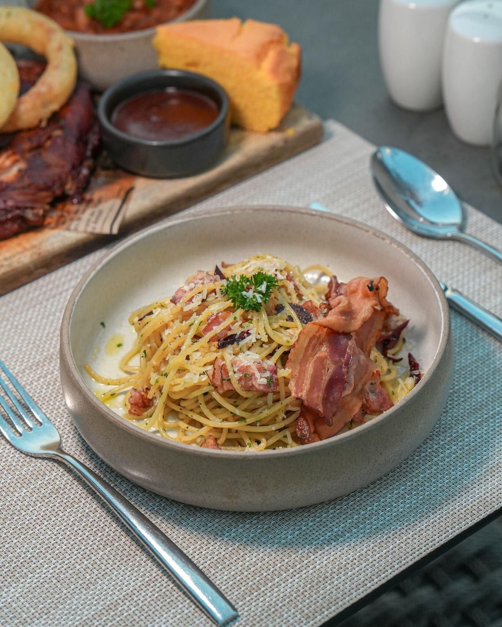 Um prato de espaguete com bacon e um enfeite, acompanhado de uma pequena tigela de molho e um pedaço de pão sobre uma mesa.