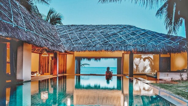 A pool area with a view of the ocean, a thatched roof structure, and a reflection in the water.