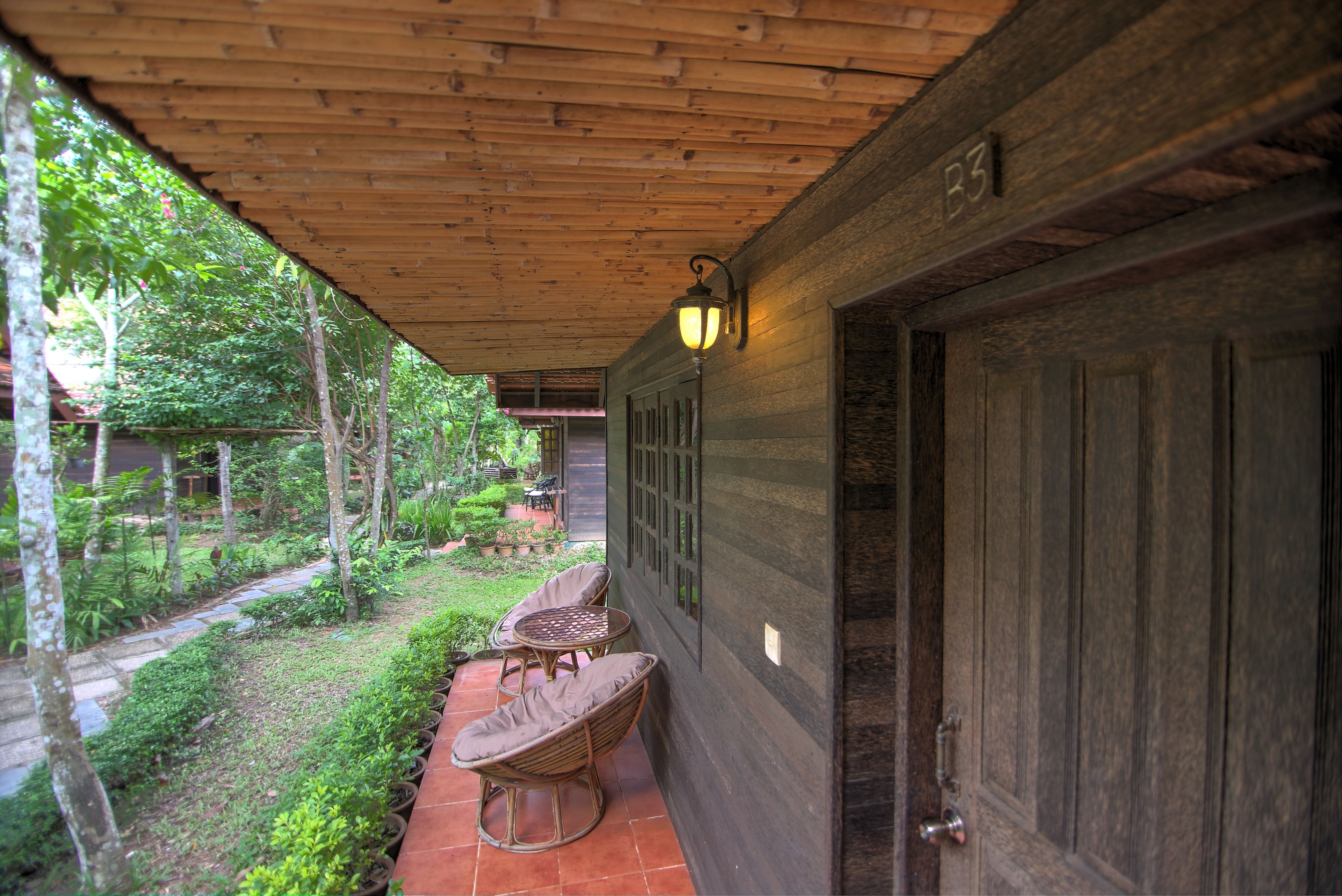 A wooden covered patio with wicker chairs and a table, surrounded by greenery.
