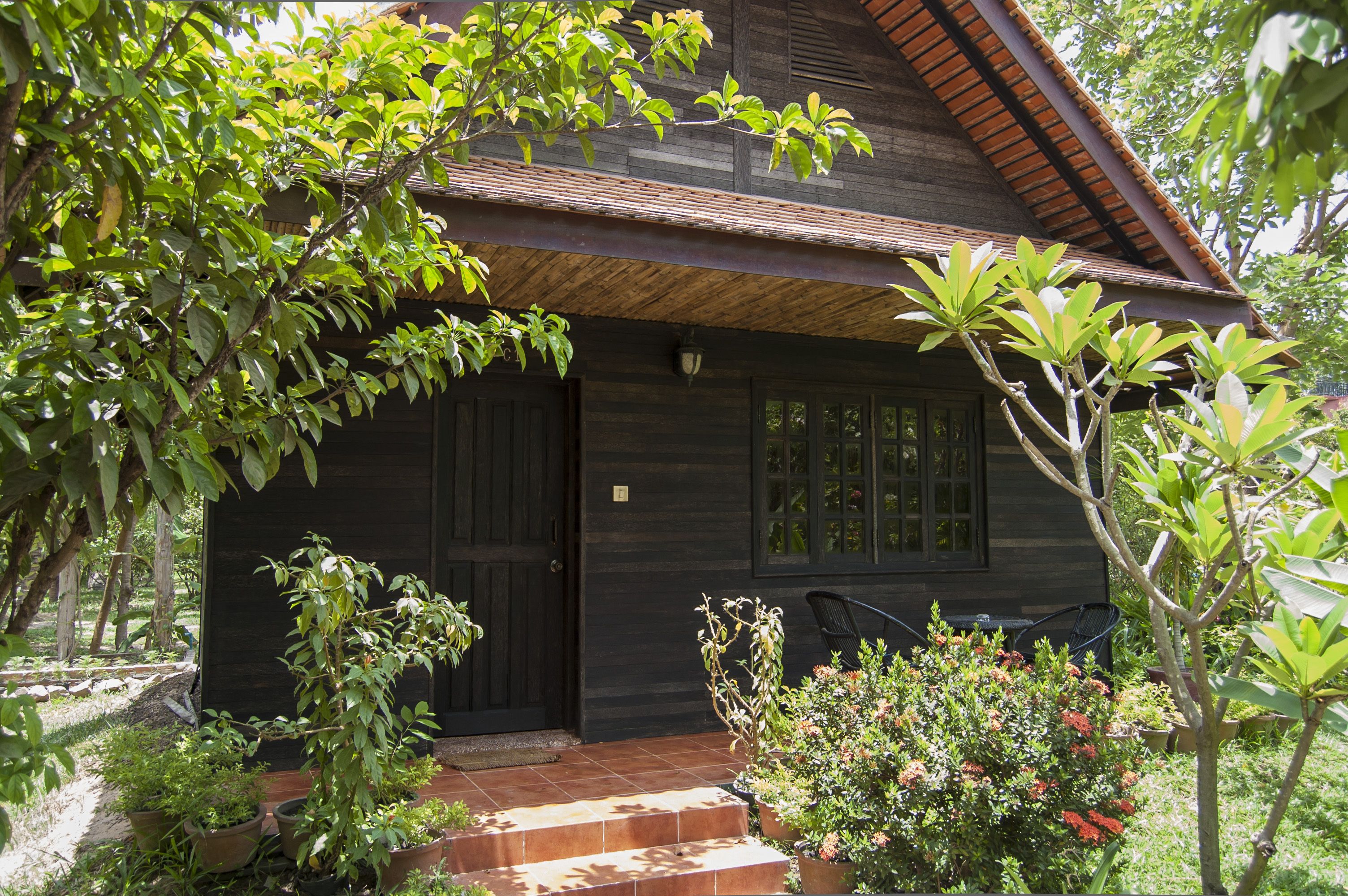 Una casa de madera con puerta oscura, ventanas y techo de tejas. Hay plantas en macetas y una silla afuera.