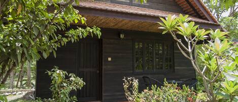 Una casa de madera con puerta oscura, ventanas y techo de tejas. Hay plantas en macetas y una silla afuera.