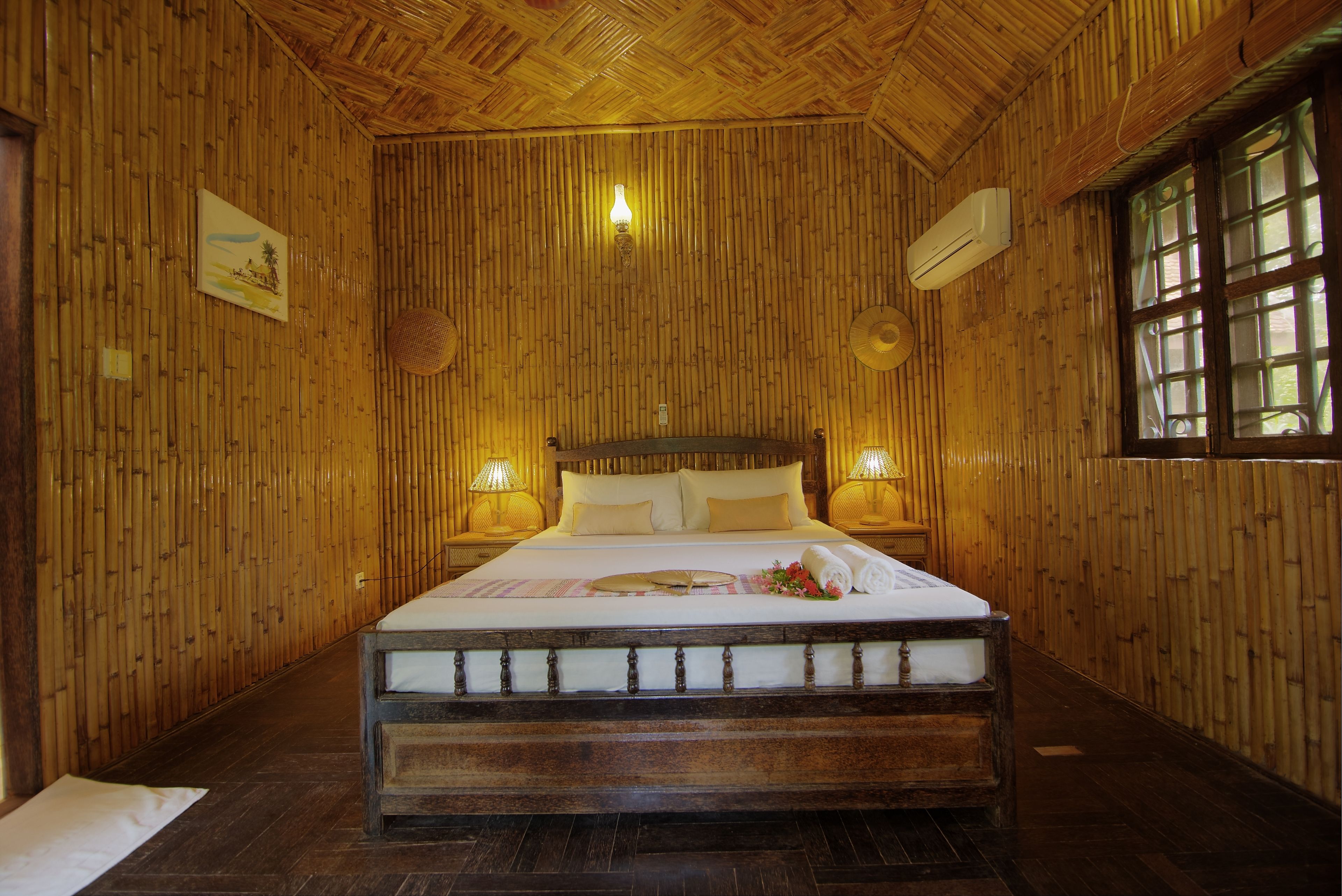Een kamer met houten plafond en wanden, een bed met houten frame, twee bedlampjes, en een raam met een rasterpatroon.